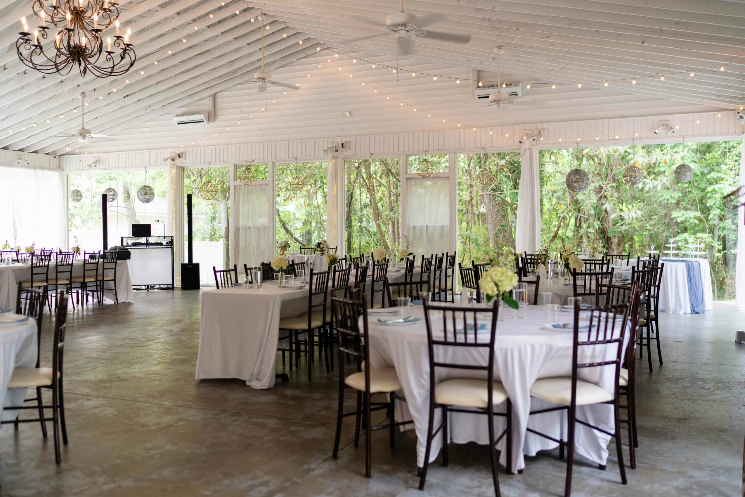 Event hall with round tables, white tablecloths, chairs, large windows, and string lights, decorated for a celebration or gathering.