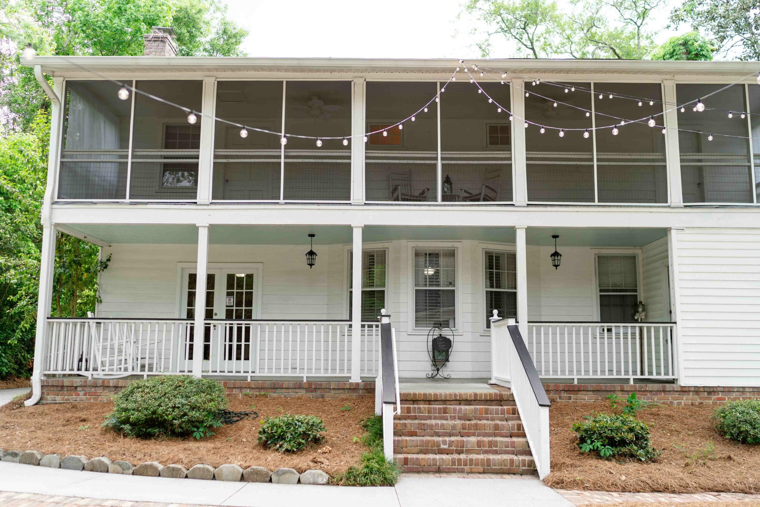 Two-story house with a porch, string lights, and a brick staircase leading to the front door, surrounded by greenery.