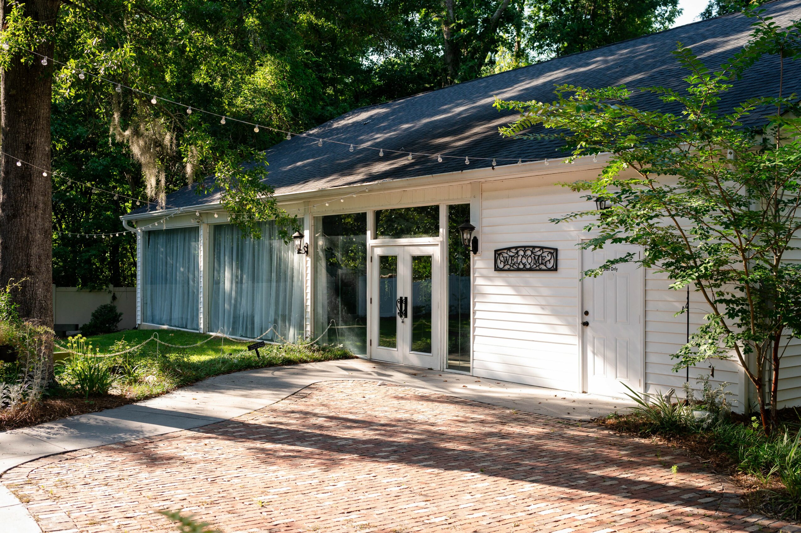 White building with glass entrance door, surrounded by trees and greenery, with a gravel pathway in front.