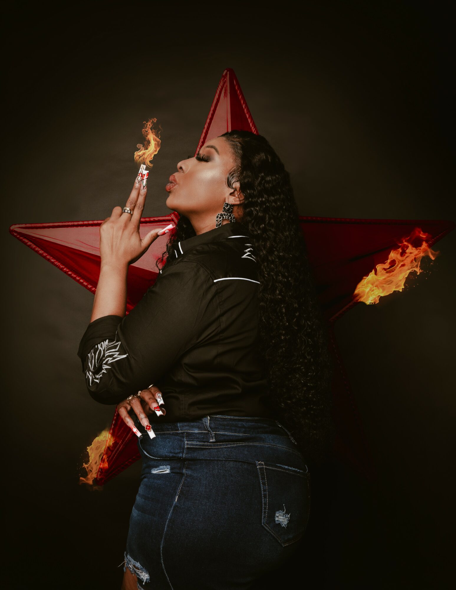 Woman with long curly hair, wearing black top and jeans, holding a lit matchstick near her face, with a red star background and flames.