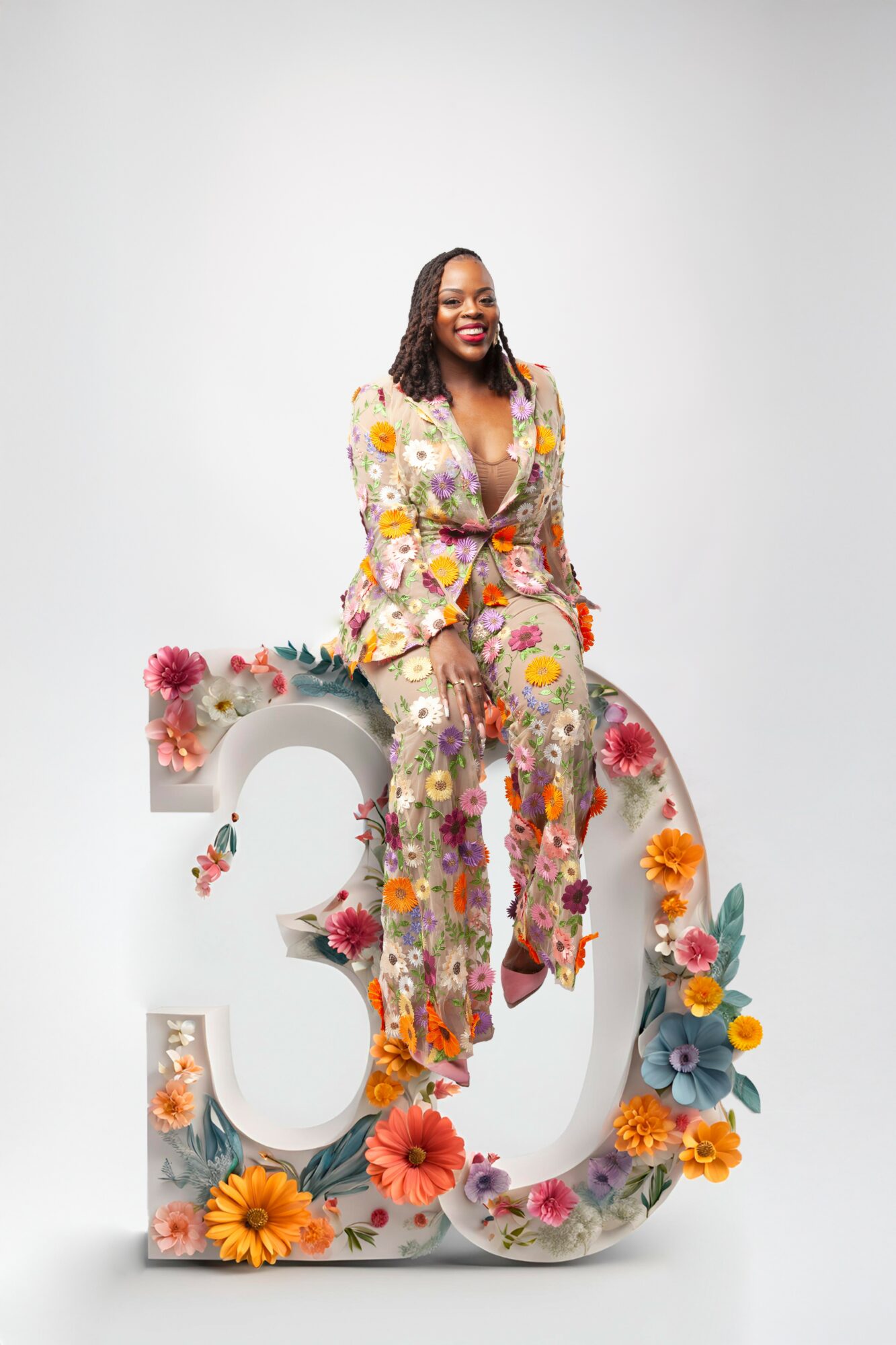Woman sitting on large decorated number 30, smiling, wearing floral outfit, surrounded by colorful flowers.