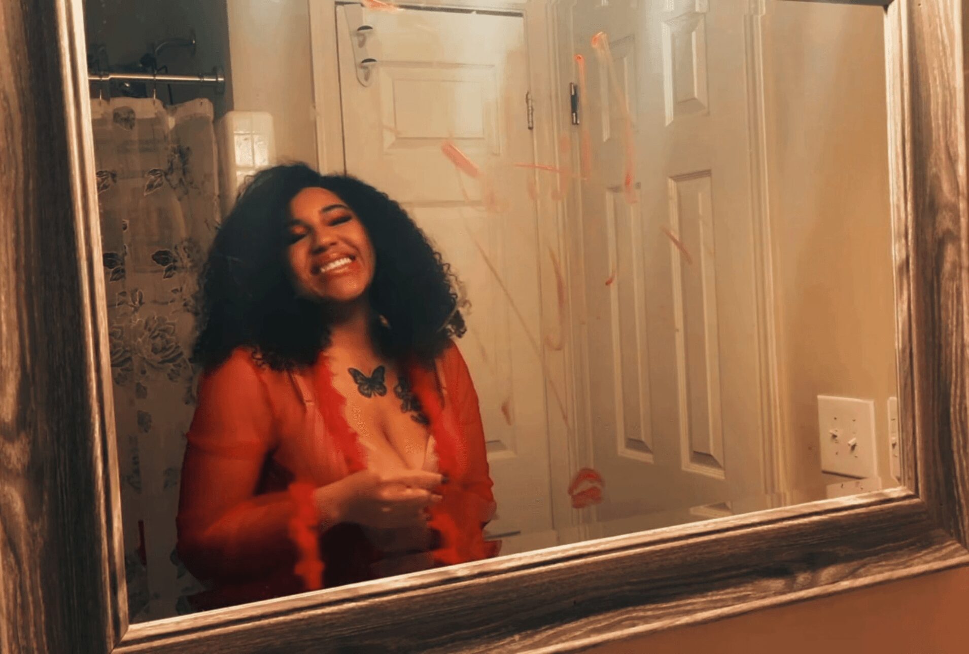 Woman with curly hair smiling in front of a mirror, holding a phone, in a room with a door and light switch.