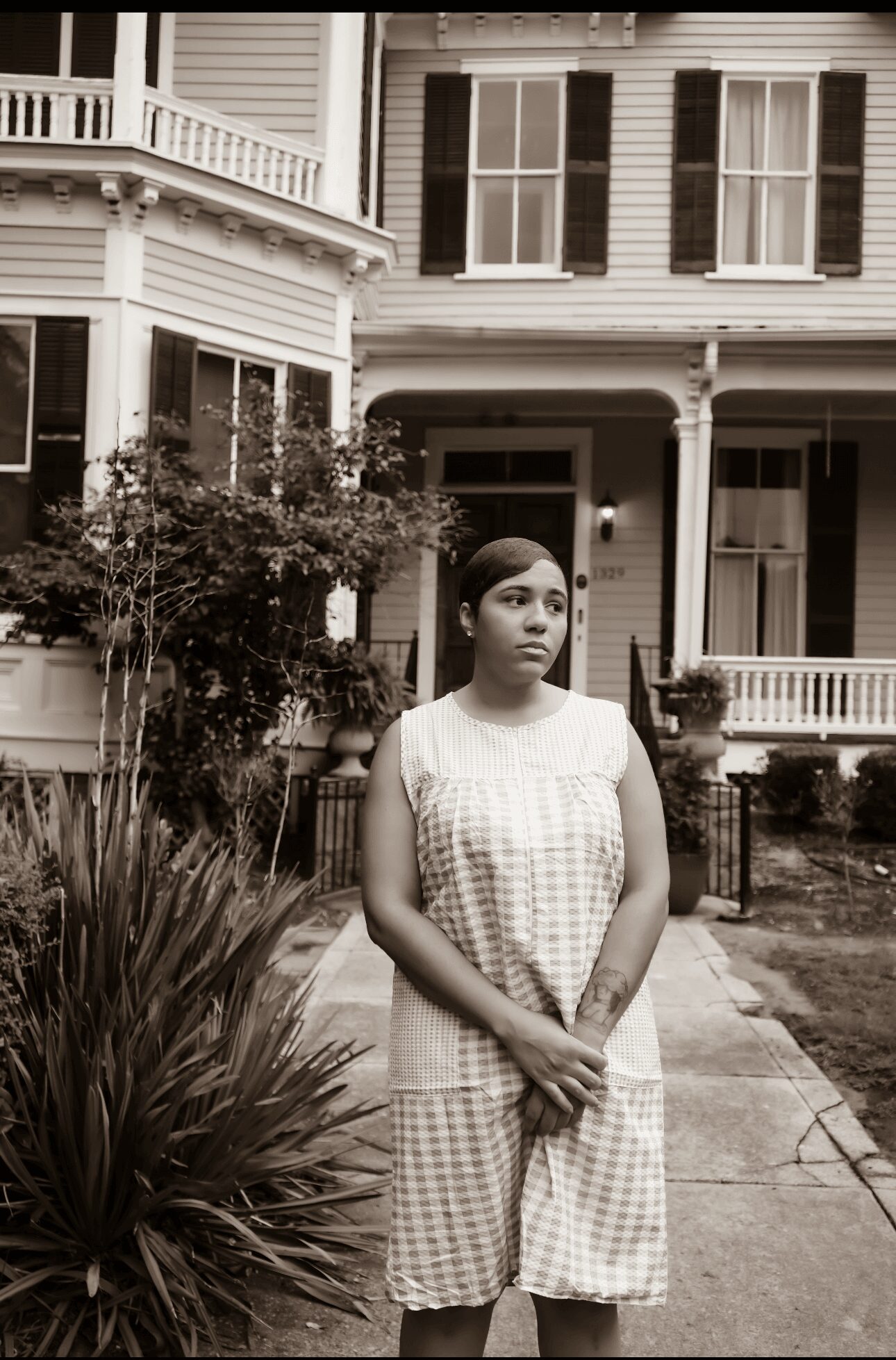 Young woman in checkered dress standing on sidewalk in front of a large house with porch and trees.