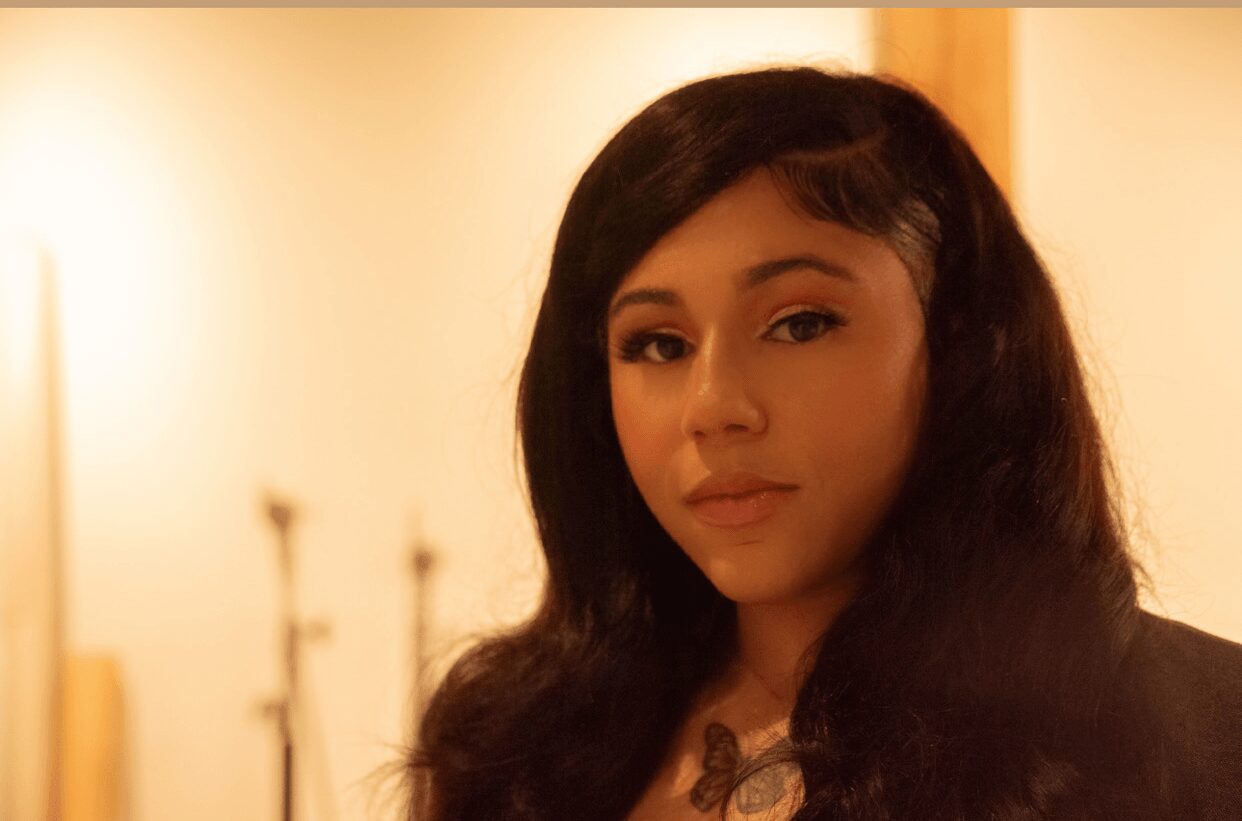 Young woman with long dark hair and makeup, looking at camera indoors with warm lighting.