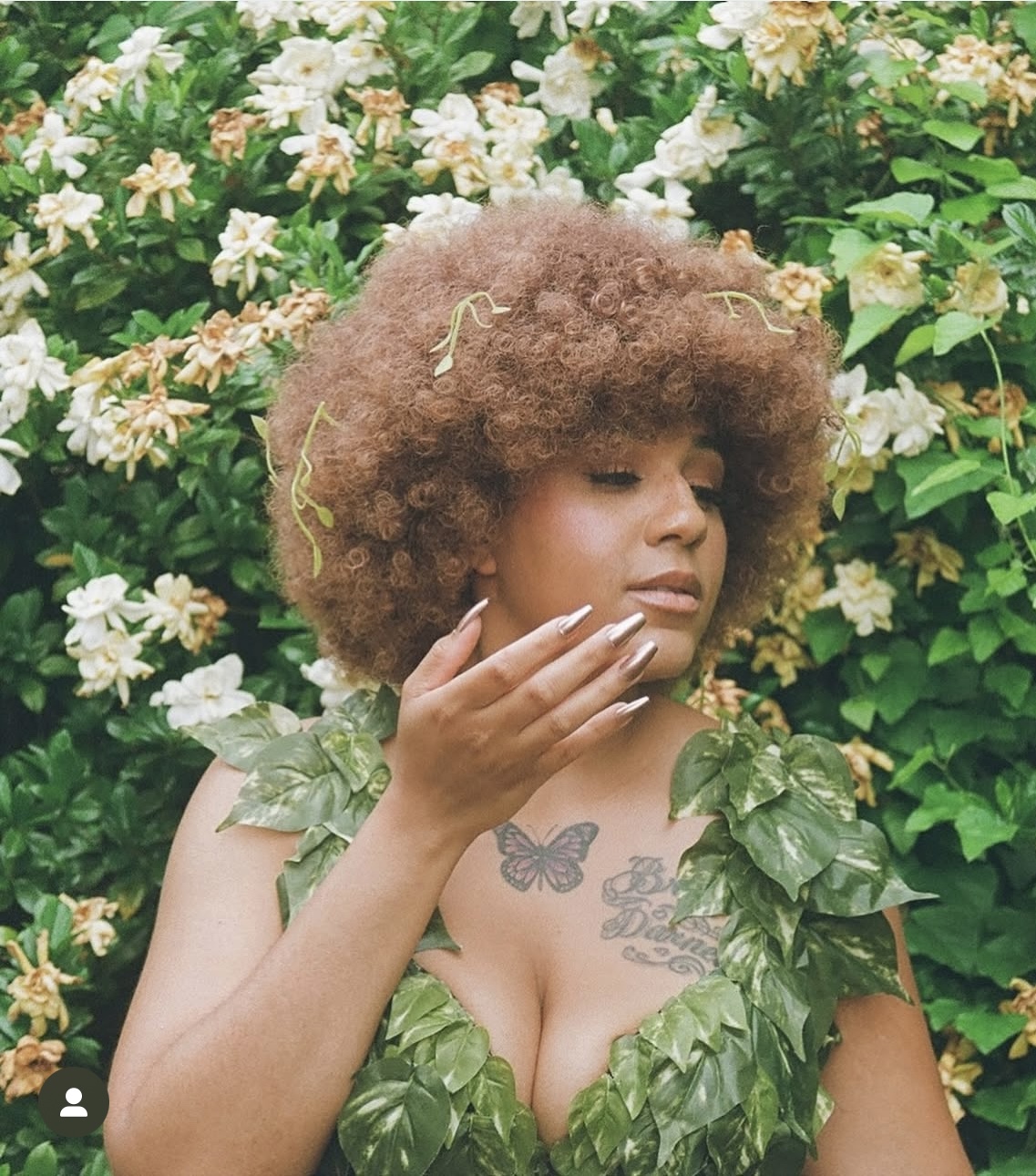 Woman with curly hair and leaf-like clothing touching her face, surrounded by white and pink flowers.