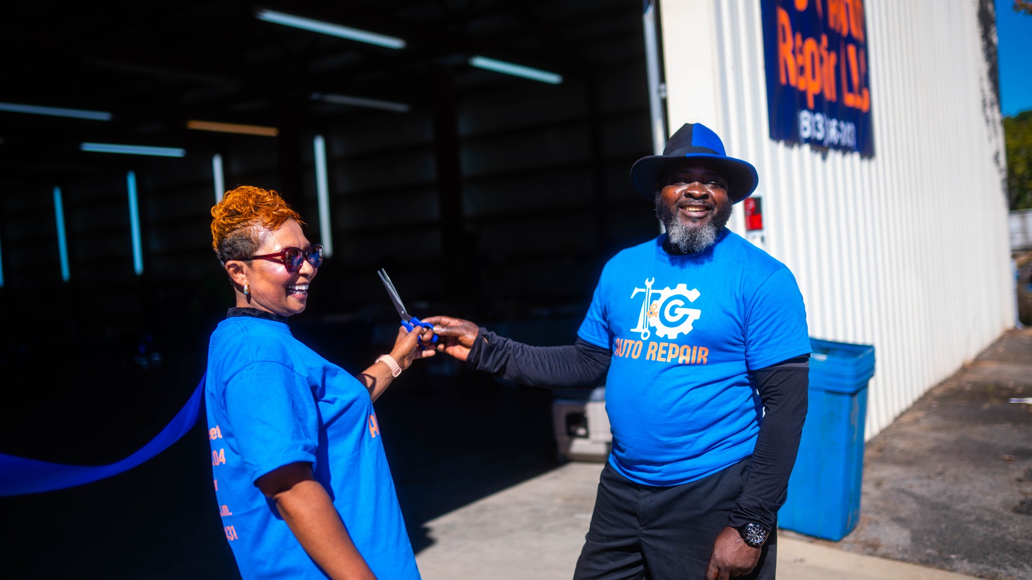 Two people exchanging a handshake outdoors, both wearing blue shirts, one with glasses and the other with a hat.