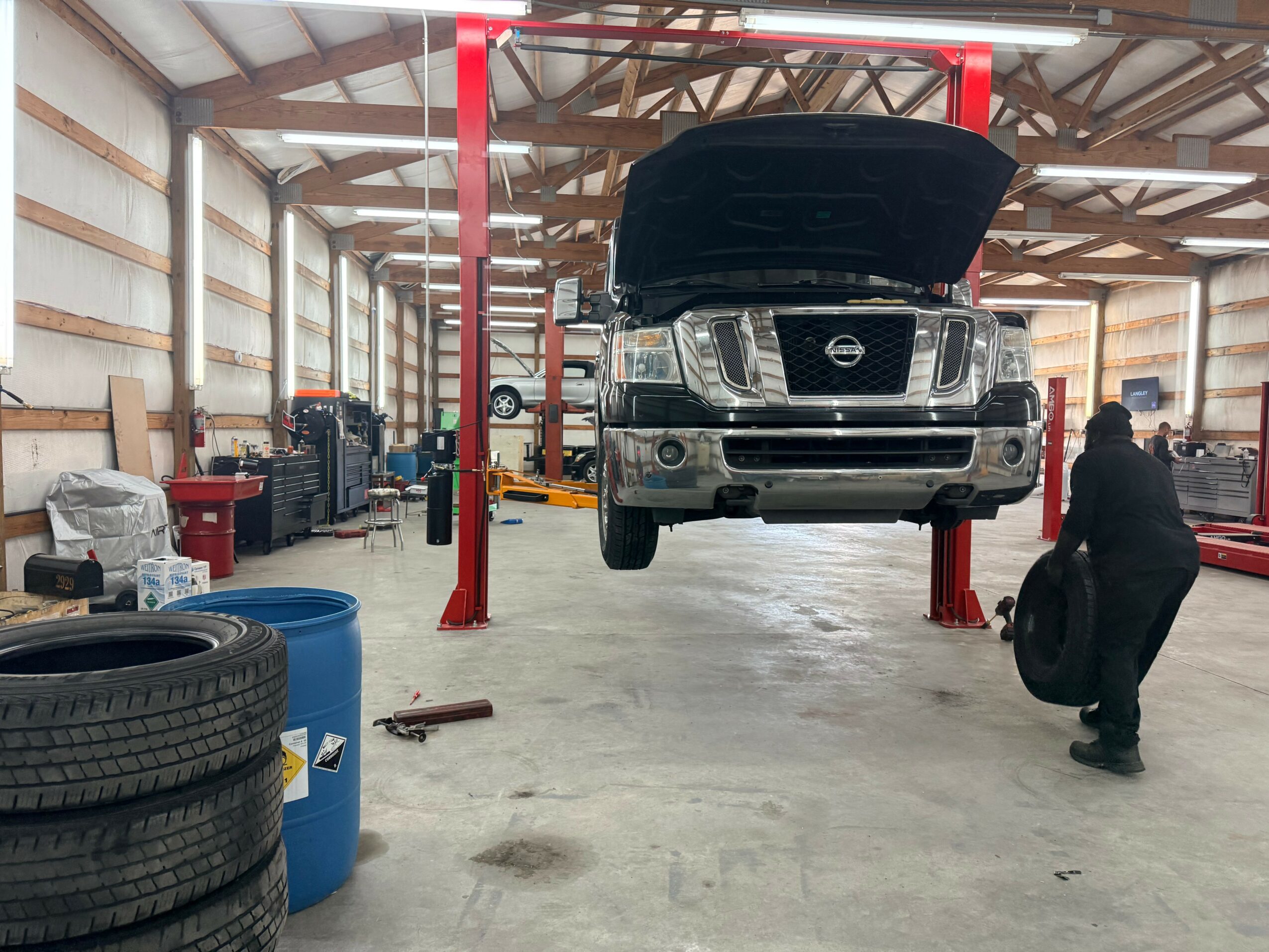 Car with open hood lifted on a hydraulic lift inside a garage, mechanic working nearby, various tools and tires around.