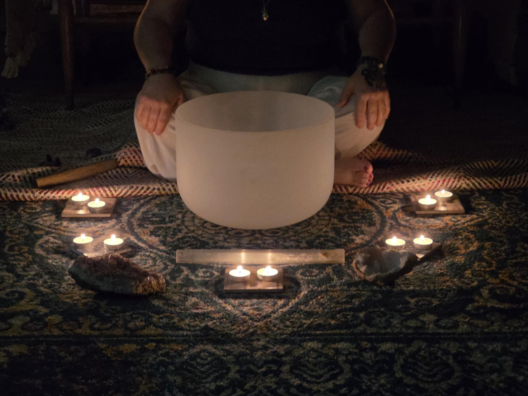 Person sitting cross-legged behind a large white singing bowl, surrounded by lit candles on a patterned rug.