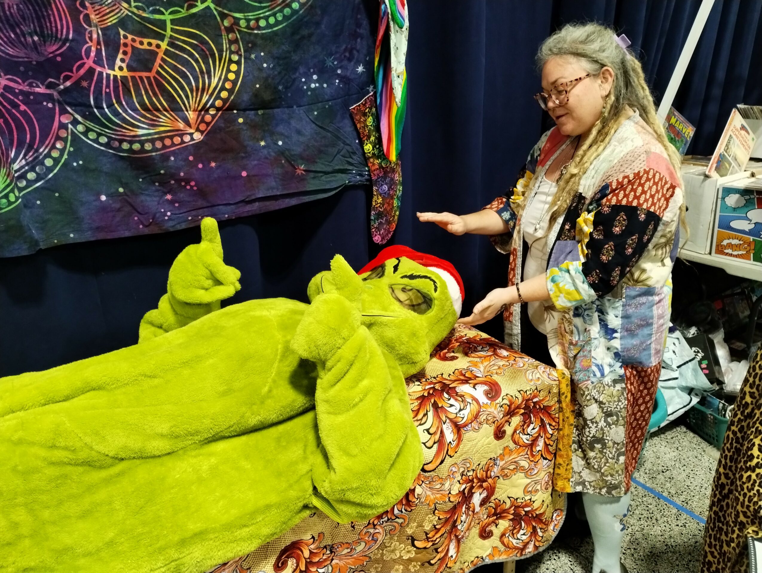 Person in a green costume lying on a table, with another person standing nearby, in front of a colorful backdrop.