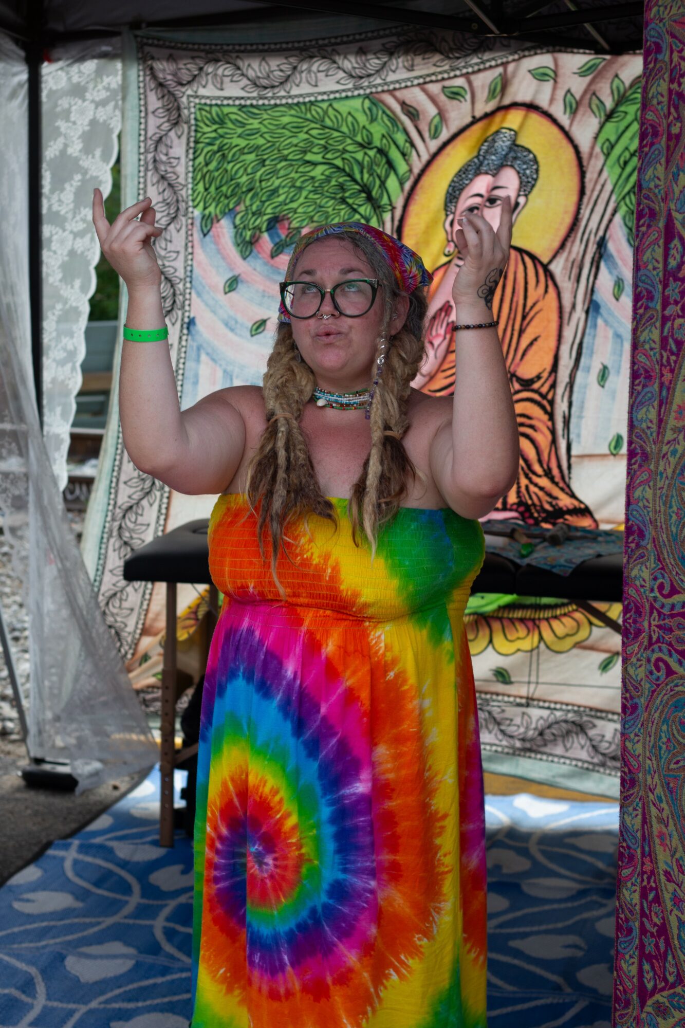 Person with long hair, glasses, wearing a rainbow tie-dye dress, raising hands, standing in front of colorful tapestry.