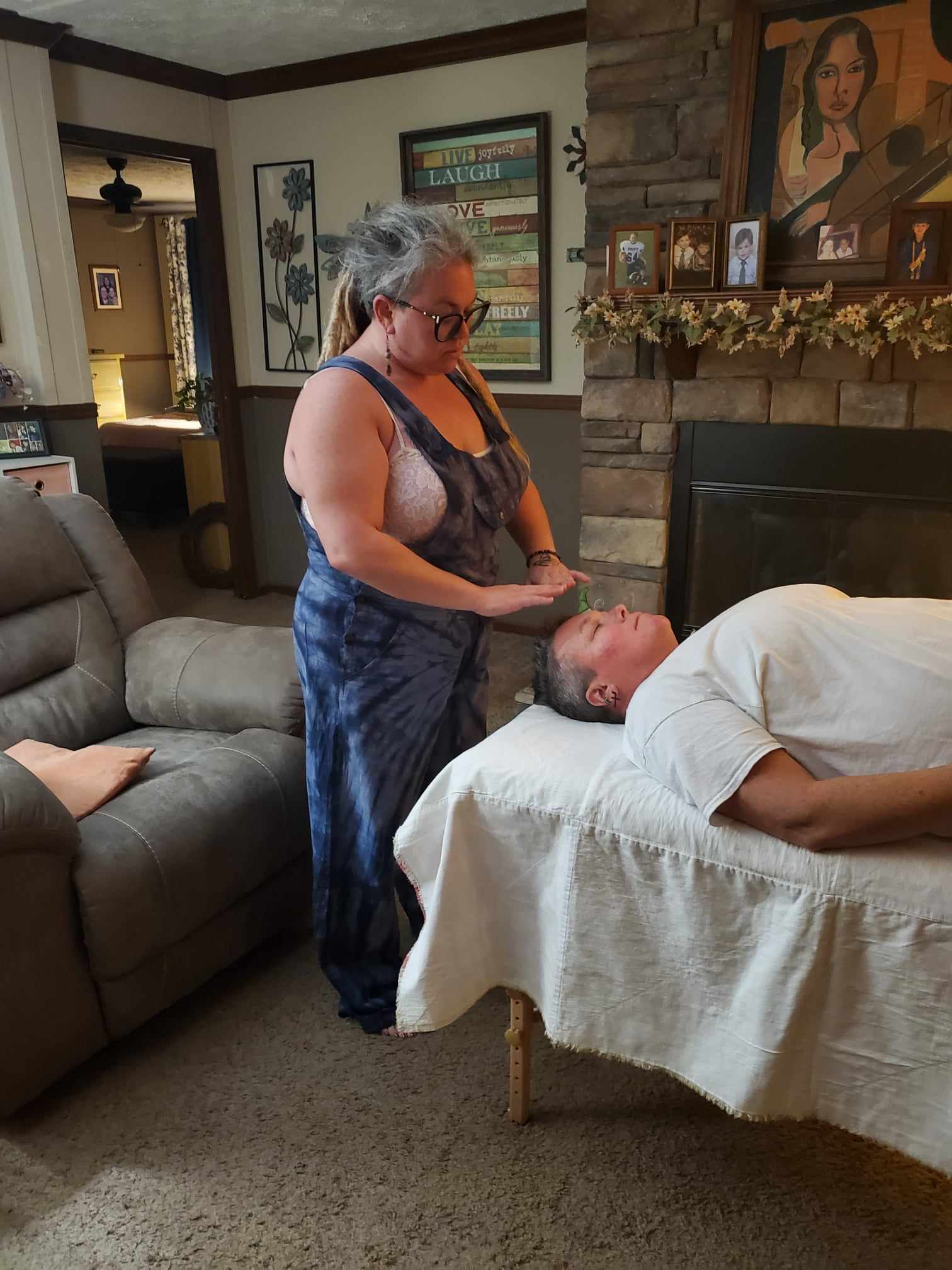 Woman with glasses and gray hair stands beside a man lying on a bed, in a living room with framed pictures and decorations.