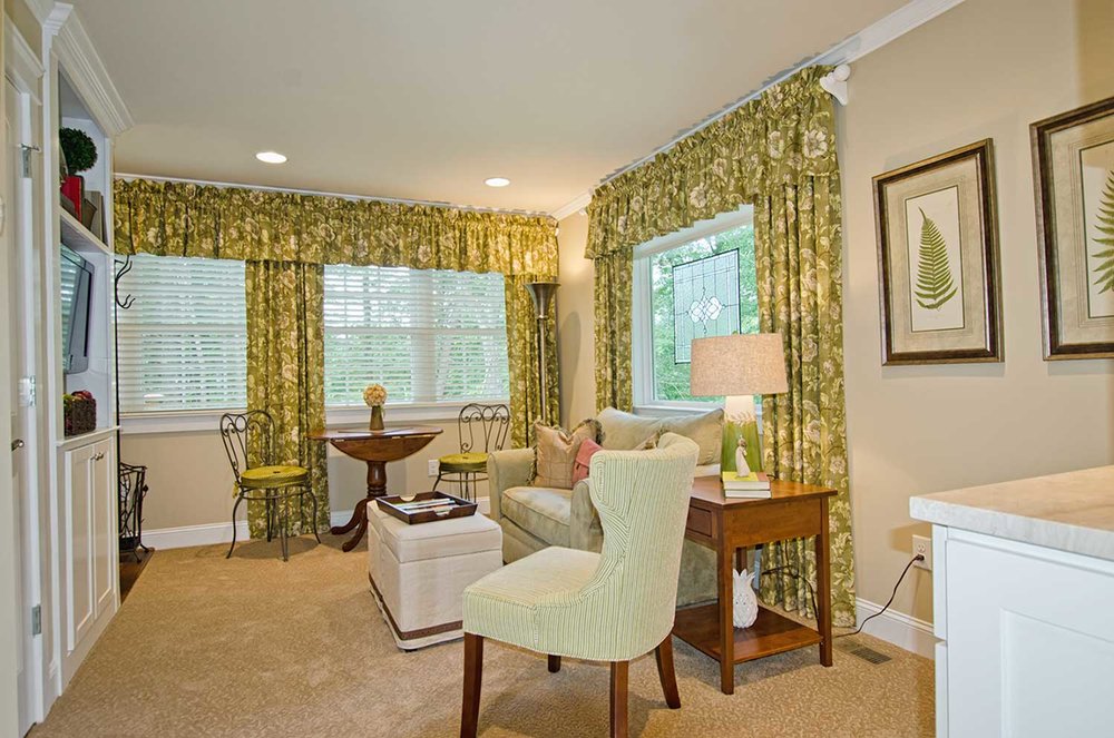 Living room with windows, green curtains, armchair, side table, lamp, and framed botanical art.