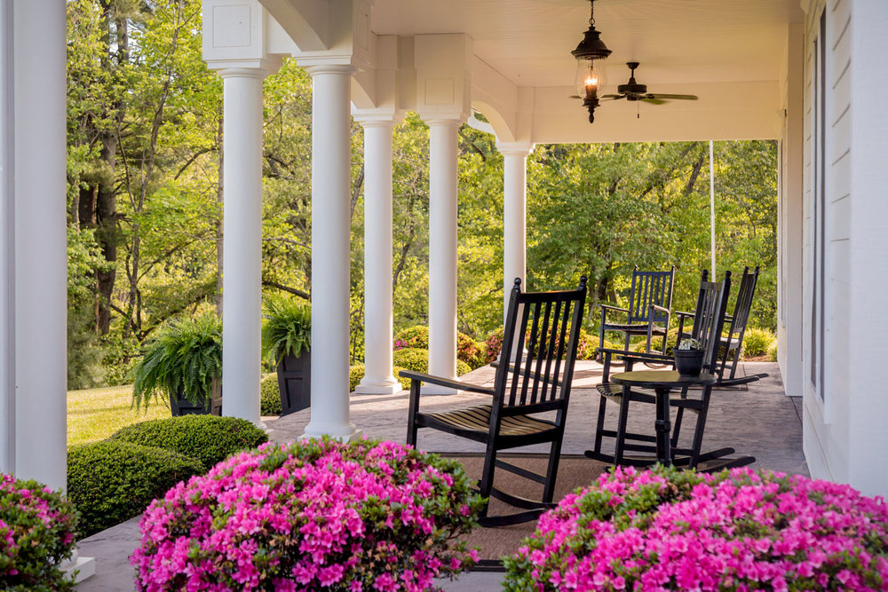 Porch with rocking chairs, white columns, pink flowers, and green trees in background.