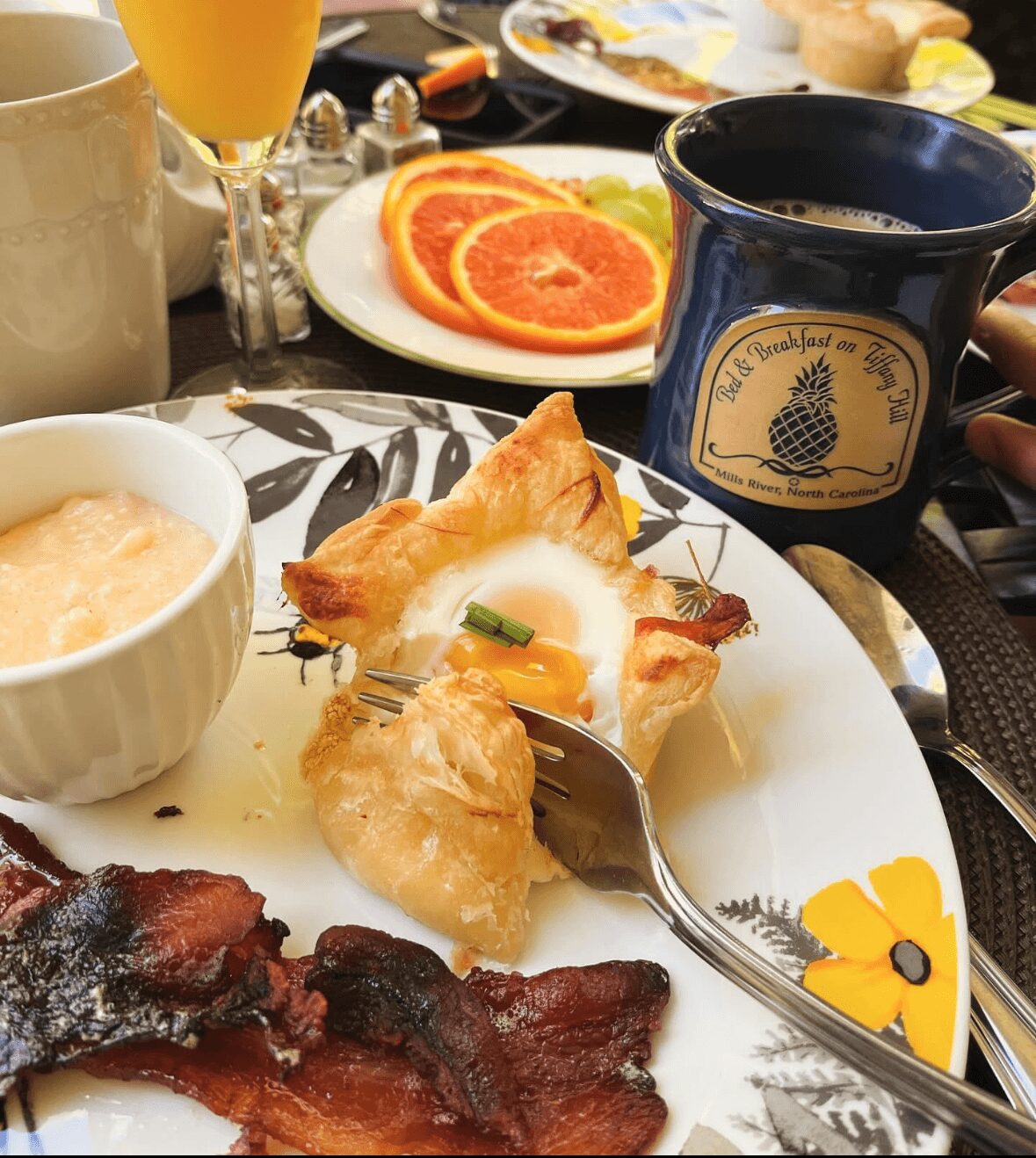 Breakfast table with orange slices, coffee, pastry with egg, bacon, and a bowl of porridge.