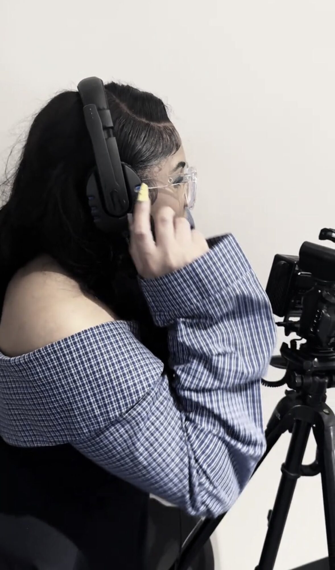 Person with long dark hair wearing headphones and glasses, adjusting headphones, in front of camera equipment.