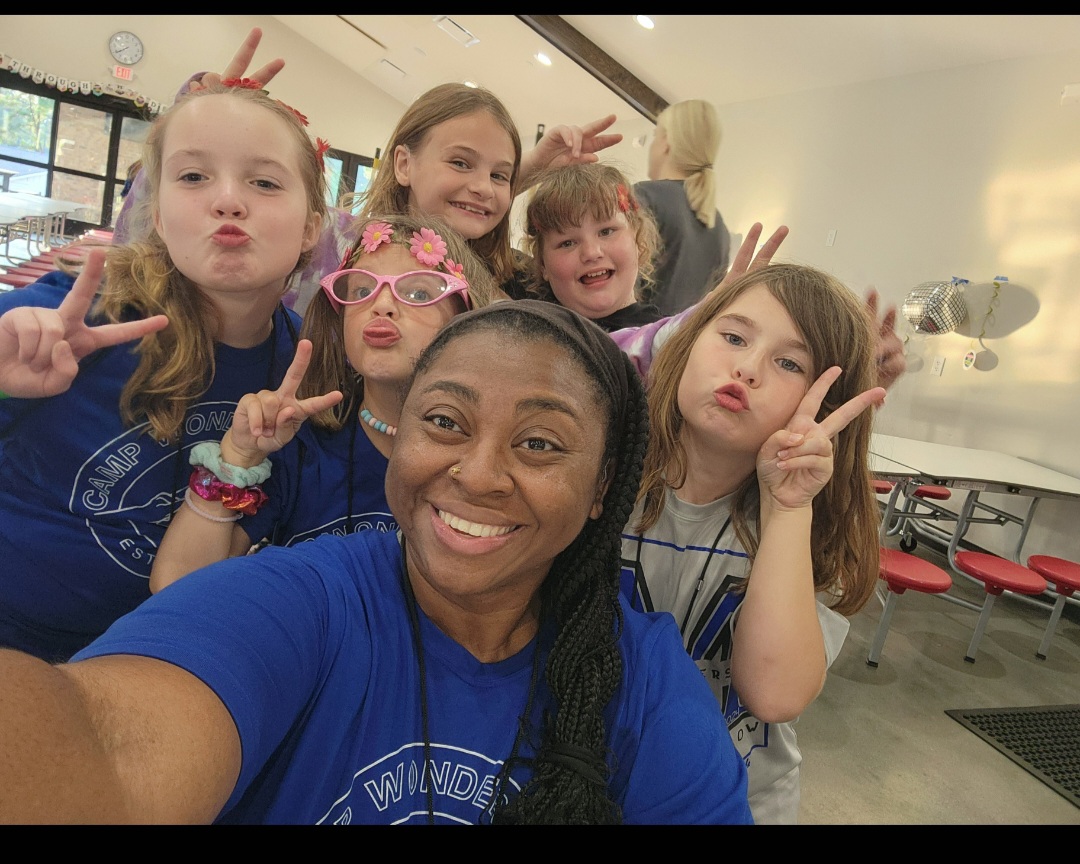 Group of children and a woman taking a selfie indoors, all making peace signs and smiling.