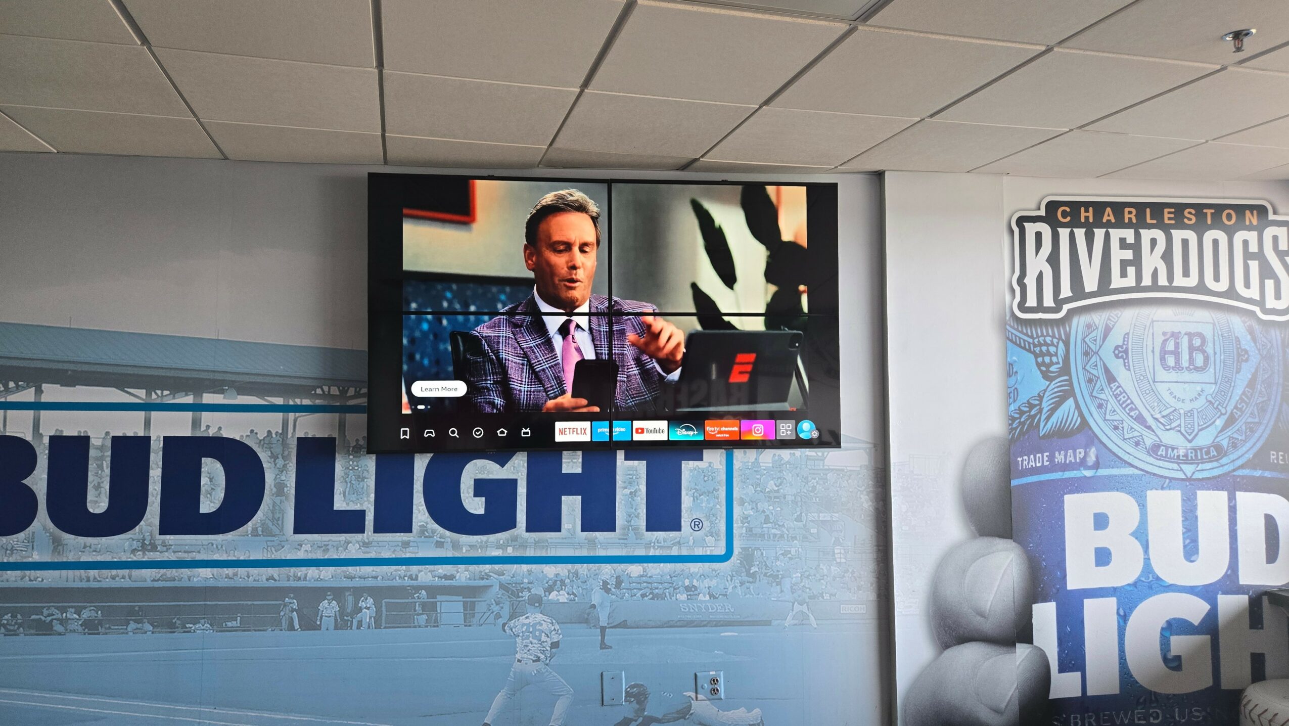 Television screen showing a man speaking in a studio with a logo and graphics behind him.