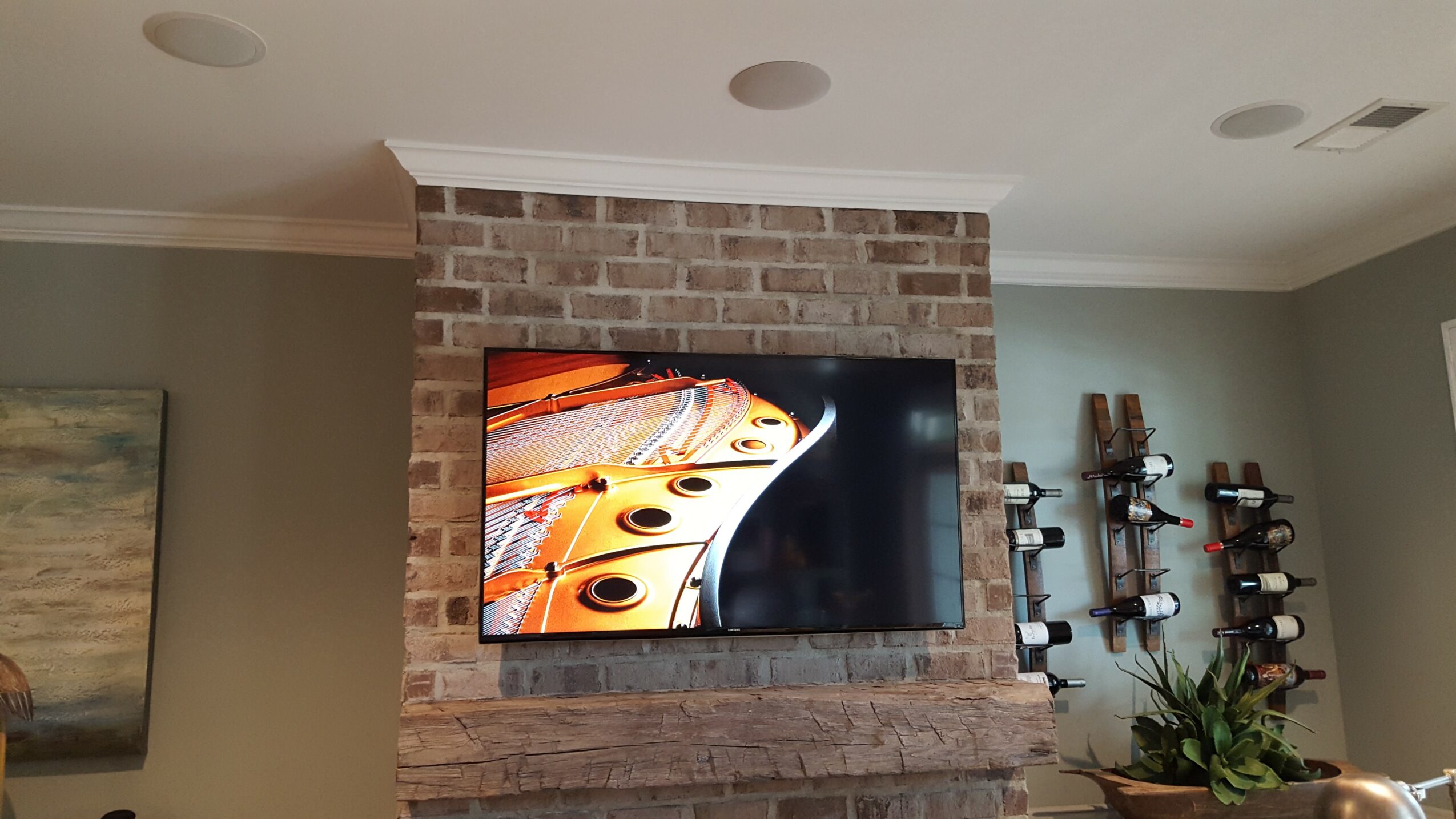 Flat-screen TV mounted on a brick wall with abstract artwork on the left and plant on the right.