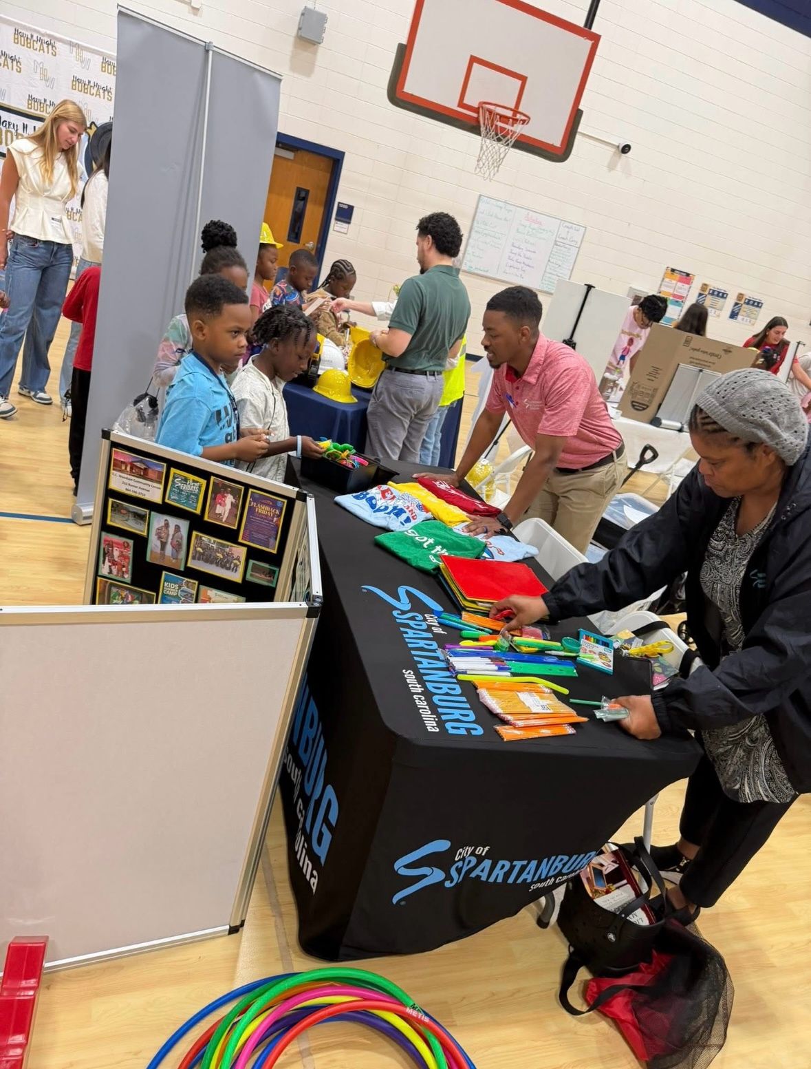 People browsing tables with colorful items in a gymnasium with basketball hoop and white walls.