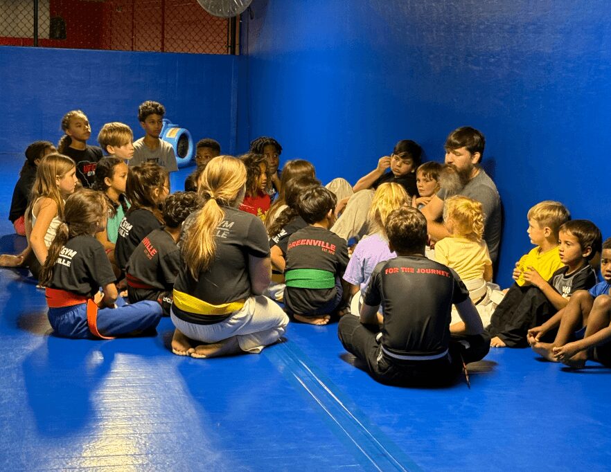 Group of children and adults sitting on a blue mat in a martial arts training room, listening to an instructor.