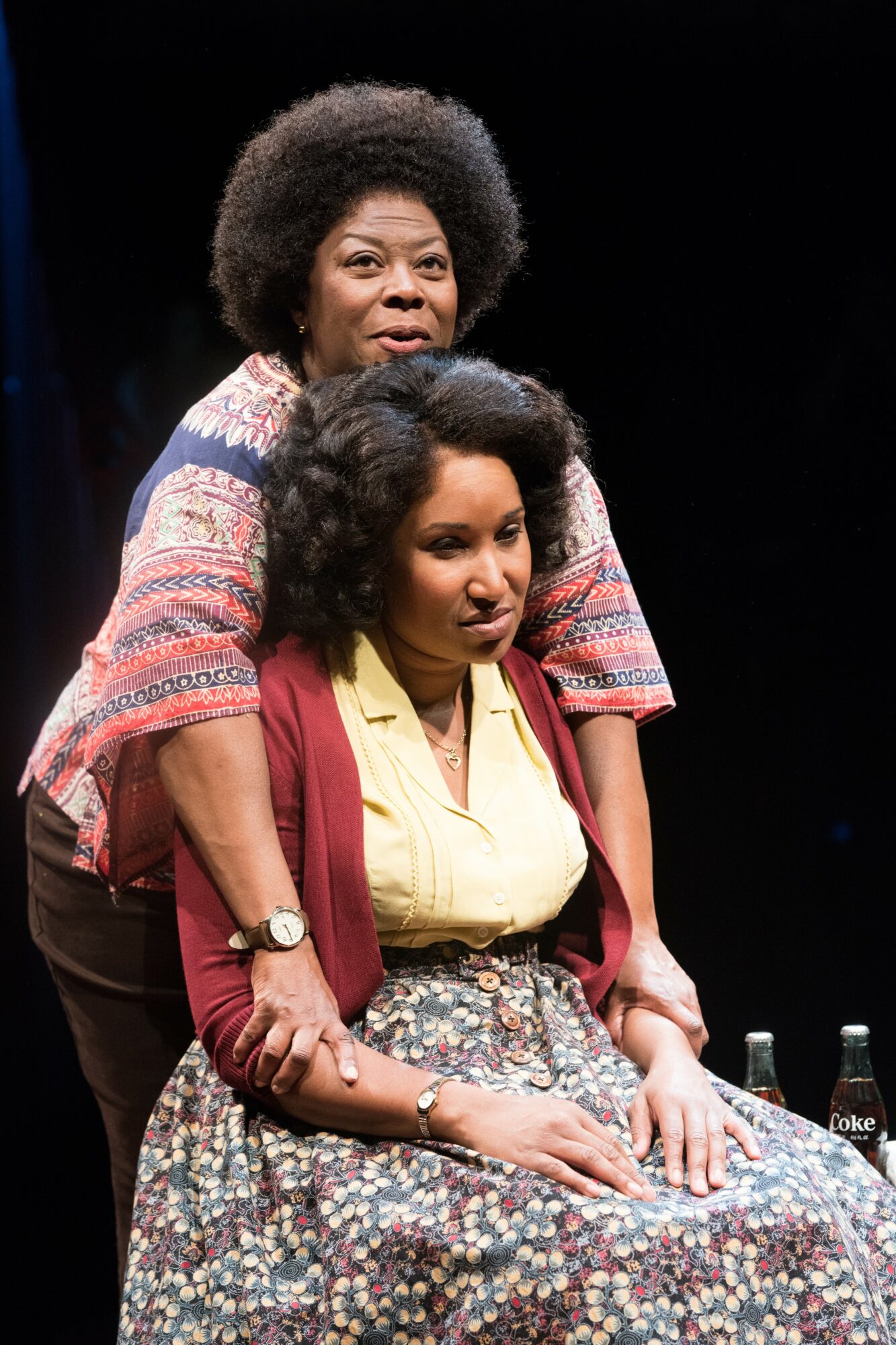 Two women, one standing behind the other, on stage with a dark background, wearing colorful clothing and jewelry.