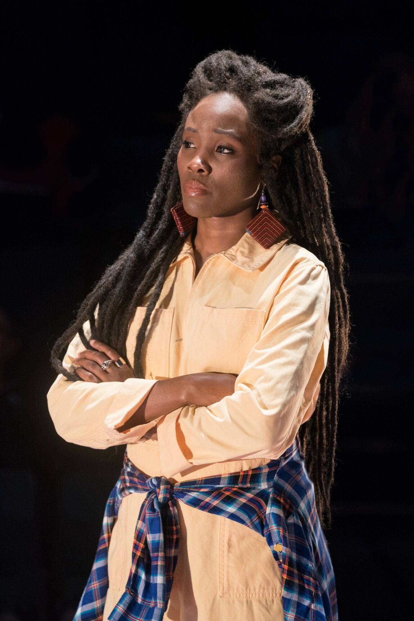 Young woman with long dreadlocks, wearing a light-colored shirt and plaid shirt tied around waist, standing with arms crossed.