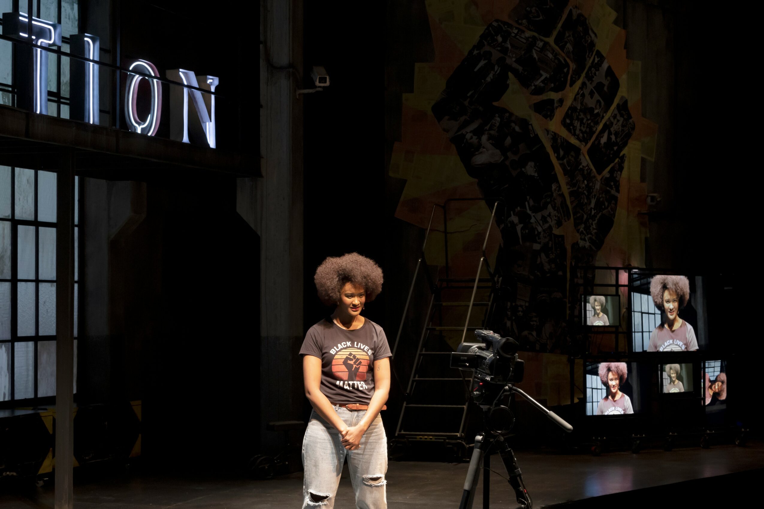 Person with curly hair standing on stage with a microphone and camera, in front of screens showing their image.