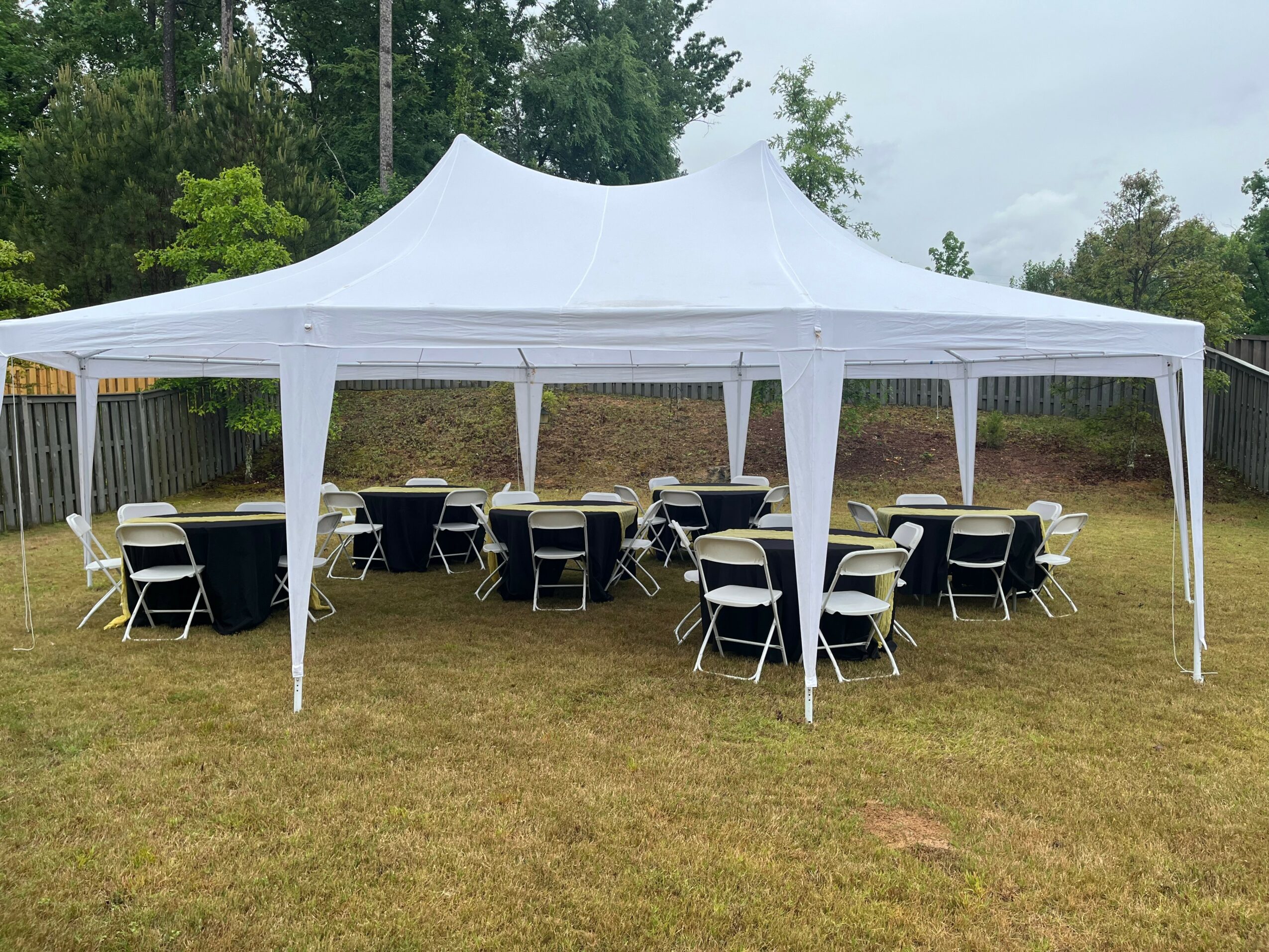 White outdoor event tent with tables and chairs on grass, surrounded by trees and a fence.