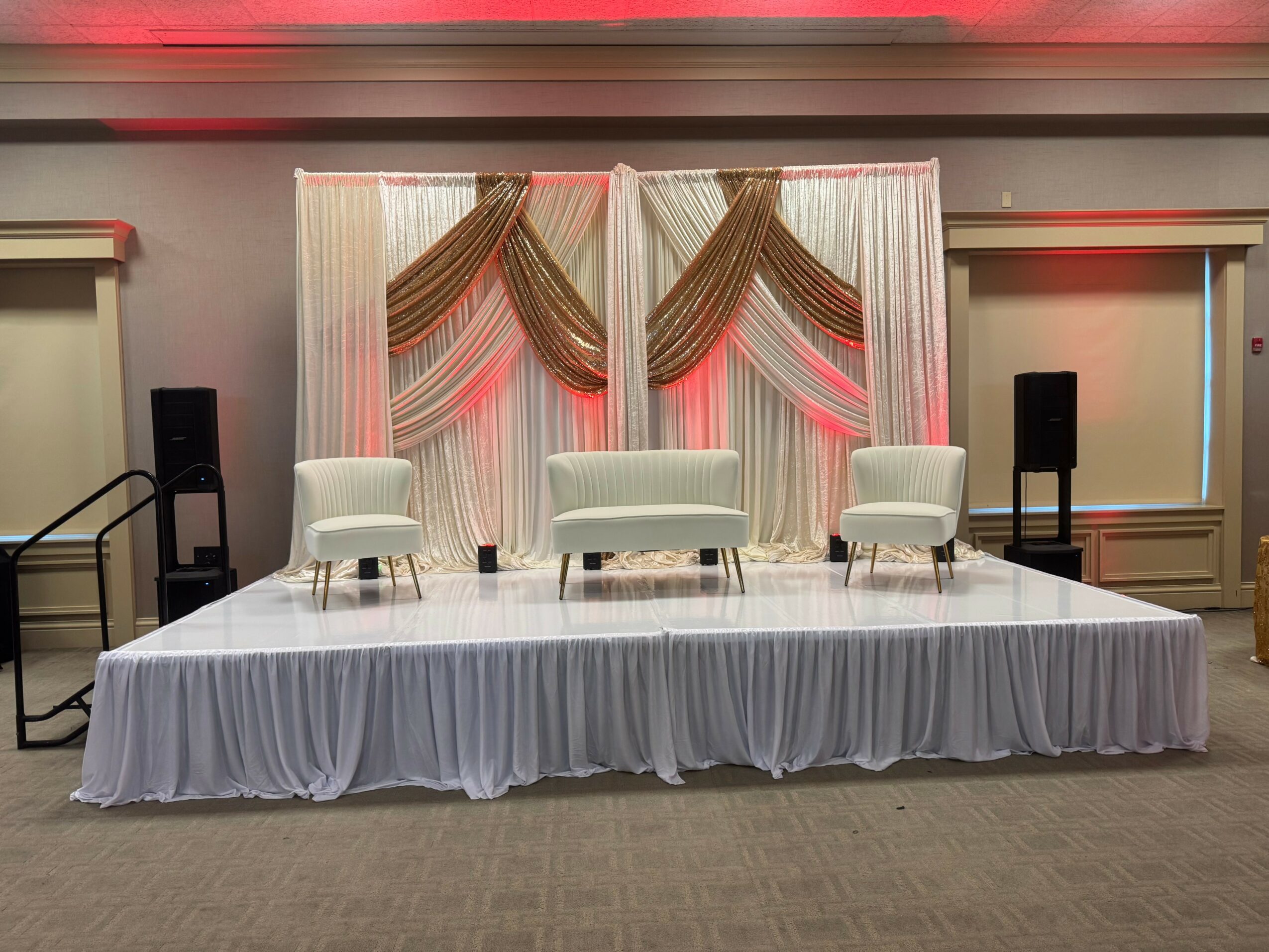 Stage with white draped backdrop, three white chairs, and two black speakers, in a decorated event space.