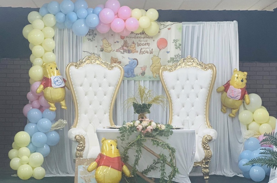 Decorative setup with two ornate white chairs, balloon arch, and Winnie the Pooh balloons, in pastel colors.