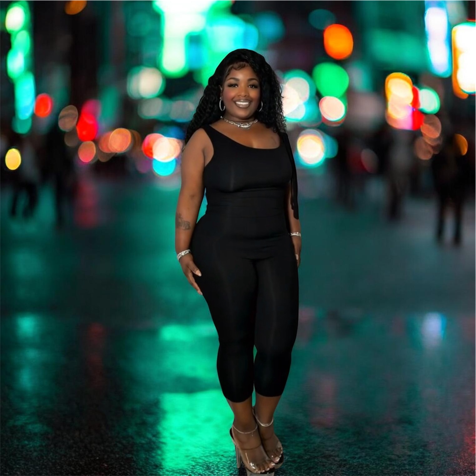 Woman standing on a city street at night with colorful blurred lights in background, smiling, wearing black outfit and heels.
