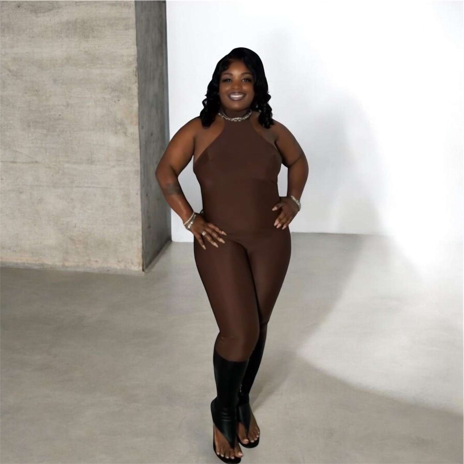 Woman in brown sleeveless jumpsuit and black boots standing indoors, smiling, with a concrete wall and white background behind her.