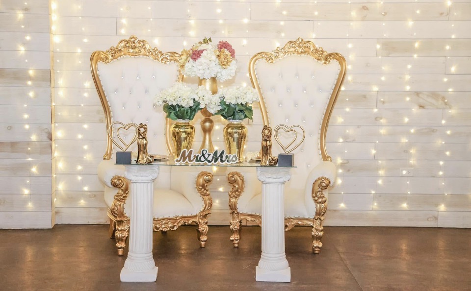 Two ornate gold and white chairs with a table in front, decorated with flowers and wedding signs, against a string light backdrop.