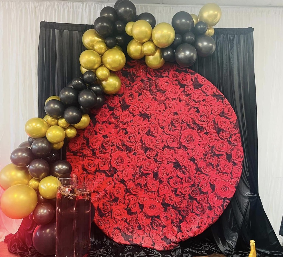 Decorative display with a large red rose circle, black and gold balloons, and a black curtain backdrop.