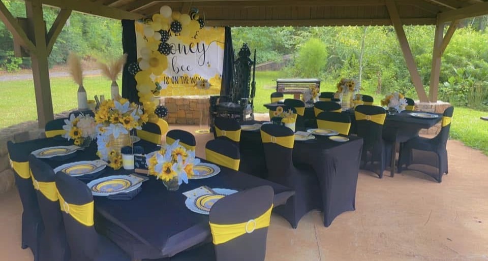 Outdoor decorated dining area with tables covered in navy blue and yellow, floral centerpieces, and a yellow backdrop with text.