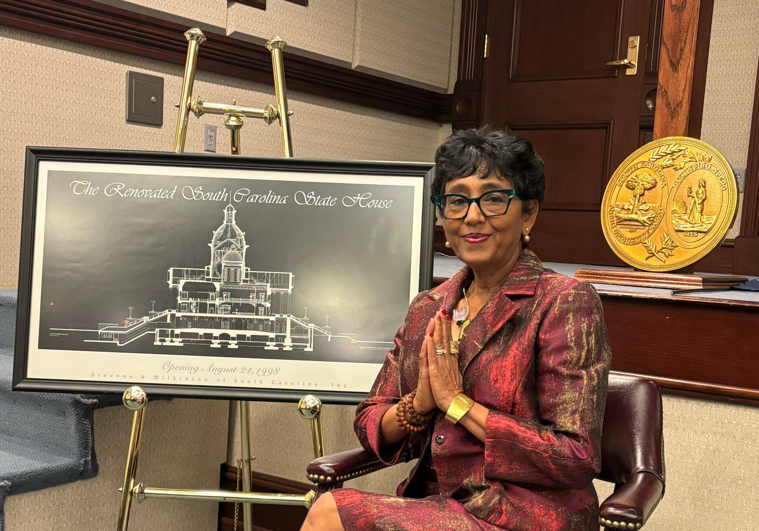 Woman with short dark hair and glasses, wearing a red patterned blazer, sitting in a chair with hands together, in a room with a framed blueprint and a gold medallion.