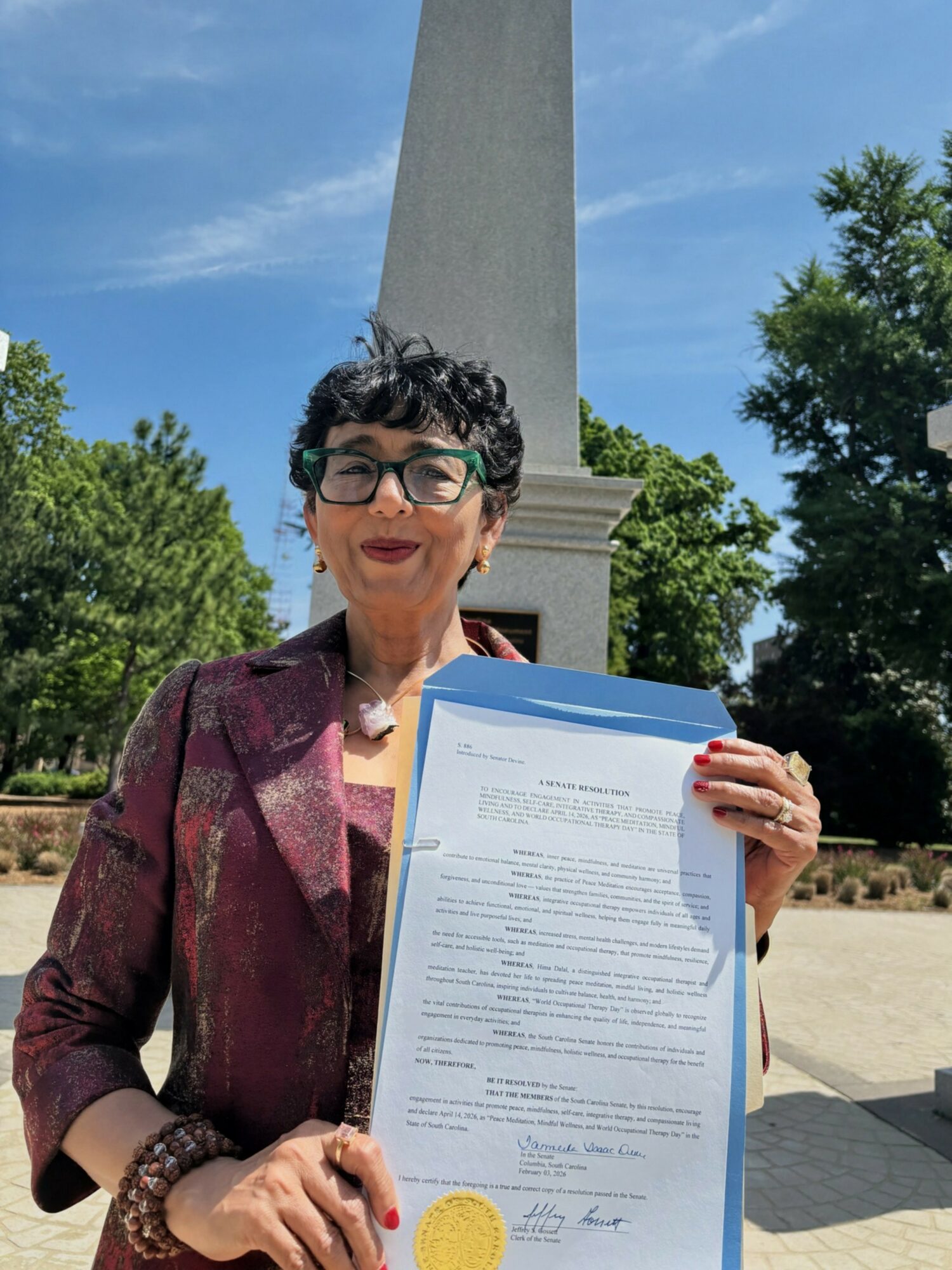 Woman with short curly hair and glasses holding a document outdoors, with trees and a monument in the background.