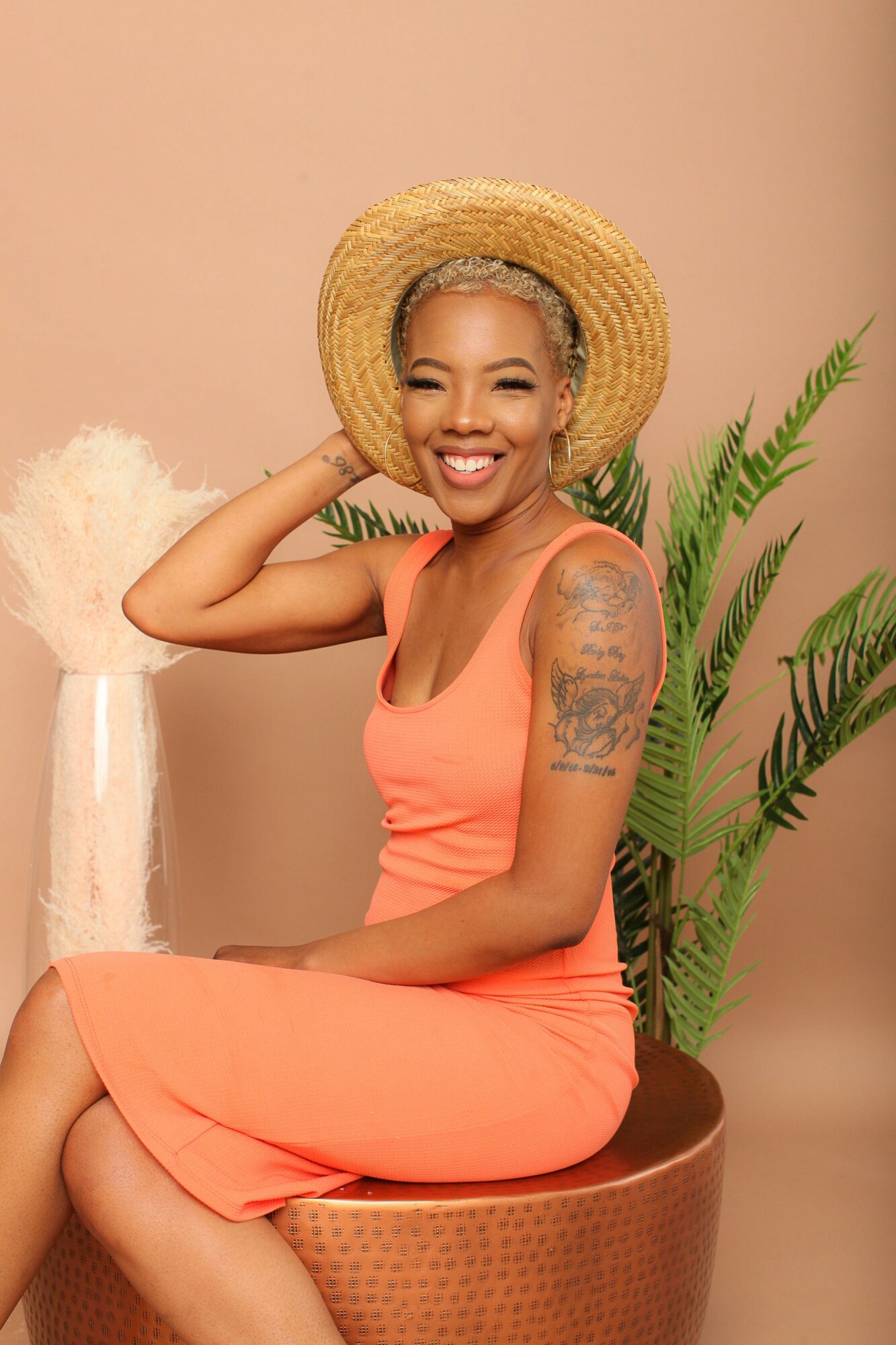Smiling woman with short gray hair wearing a straw hat and coral dress, sitting on a round stool with plants behind her.