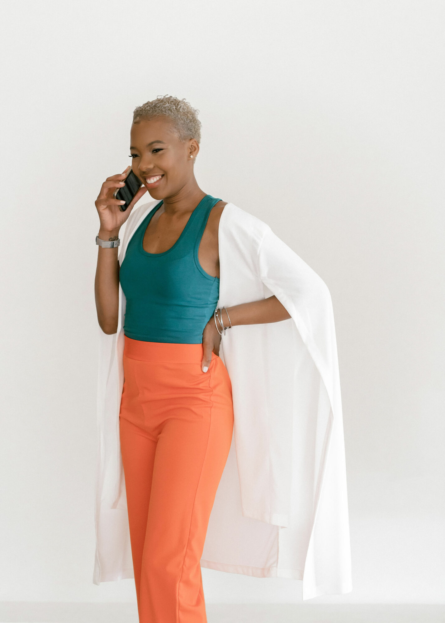 Woman with short gray hair talking on a phone, wearing a teal top, orange pants, and a white coat, smiling.
