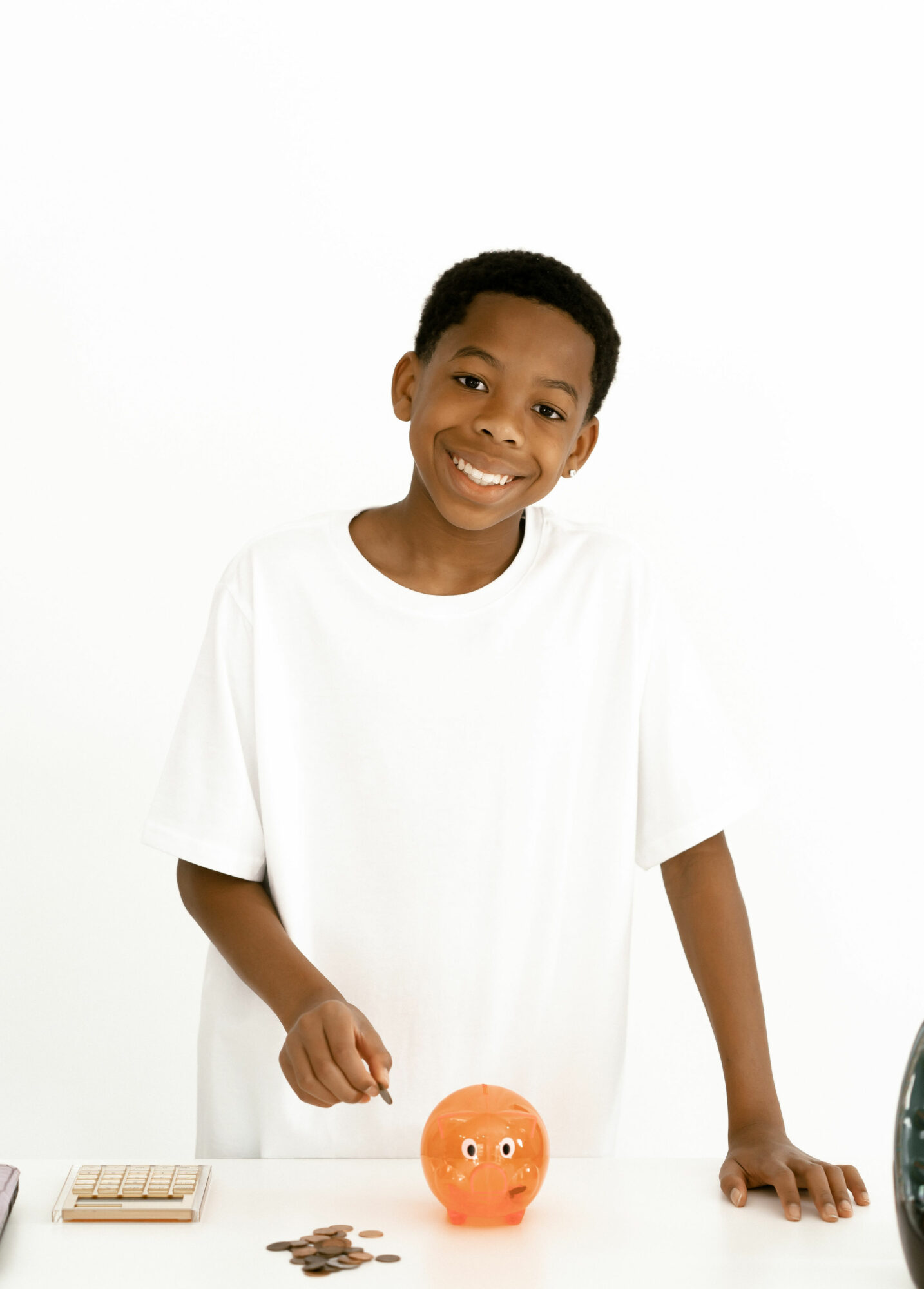 Smiling boy in a white shirt standing next to a piggy bank and coins, against a white background.