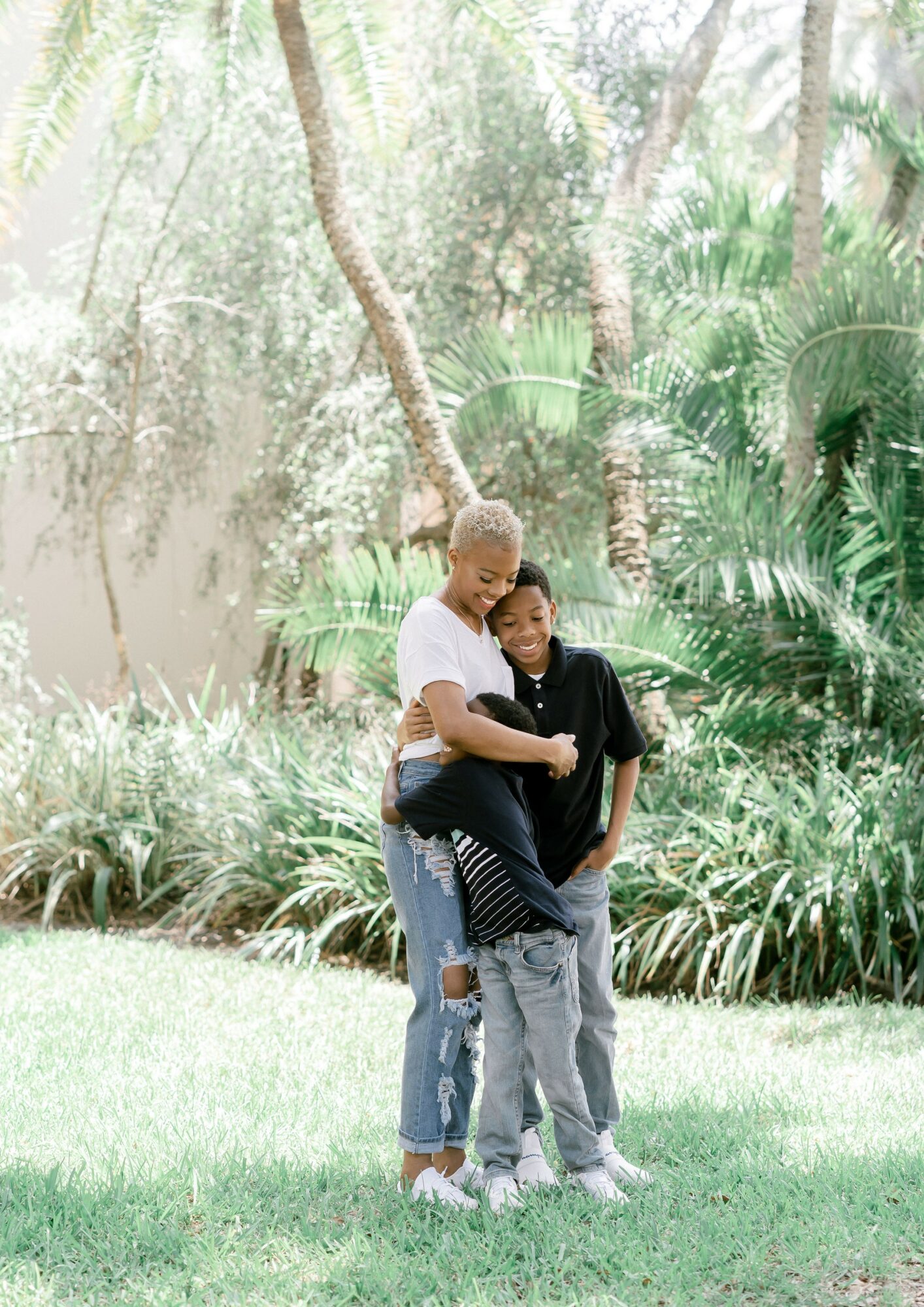 Two people standing outdoors in a lush, green park with trees and plants, embracing each other.