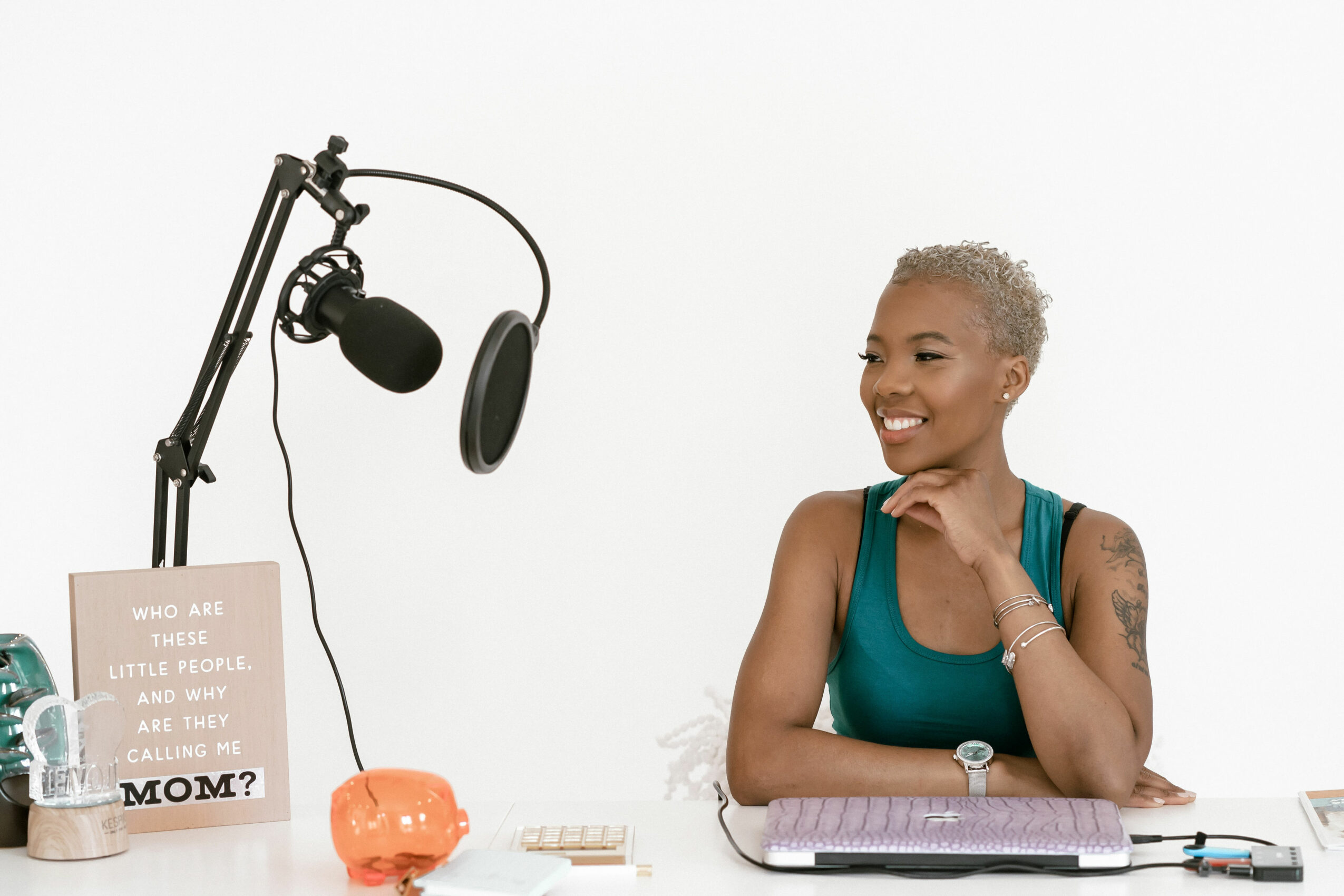Smiling woman with short gray hair sitting at a desk with recording equipment and a sign, against a white background.