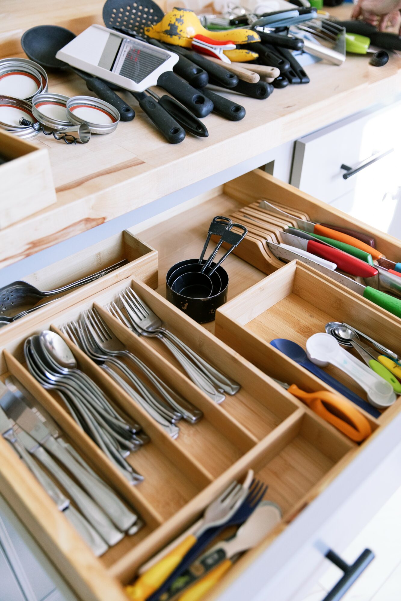 Kitchen drawer with utensils, knives, and scissors organized in wooden compartments, with tools and gadgets on the countertop above.