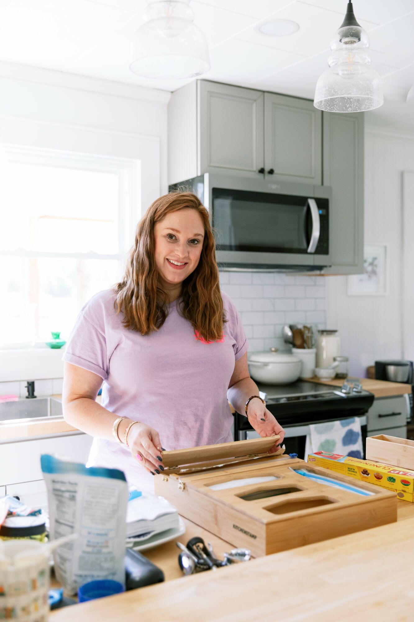 Young woman with long hair smiling in a kitchen, preparing food on a wooden cutting board, with appliances and utensils around.