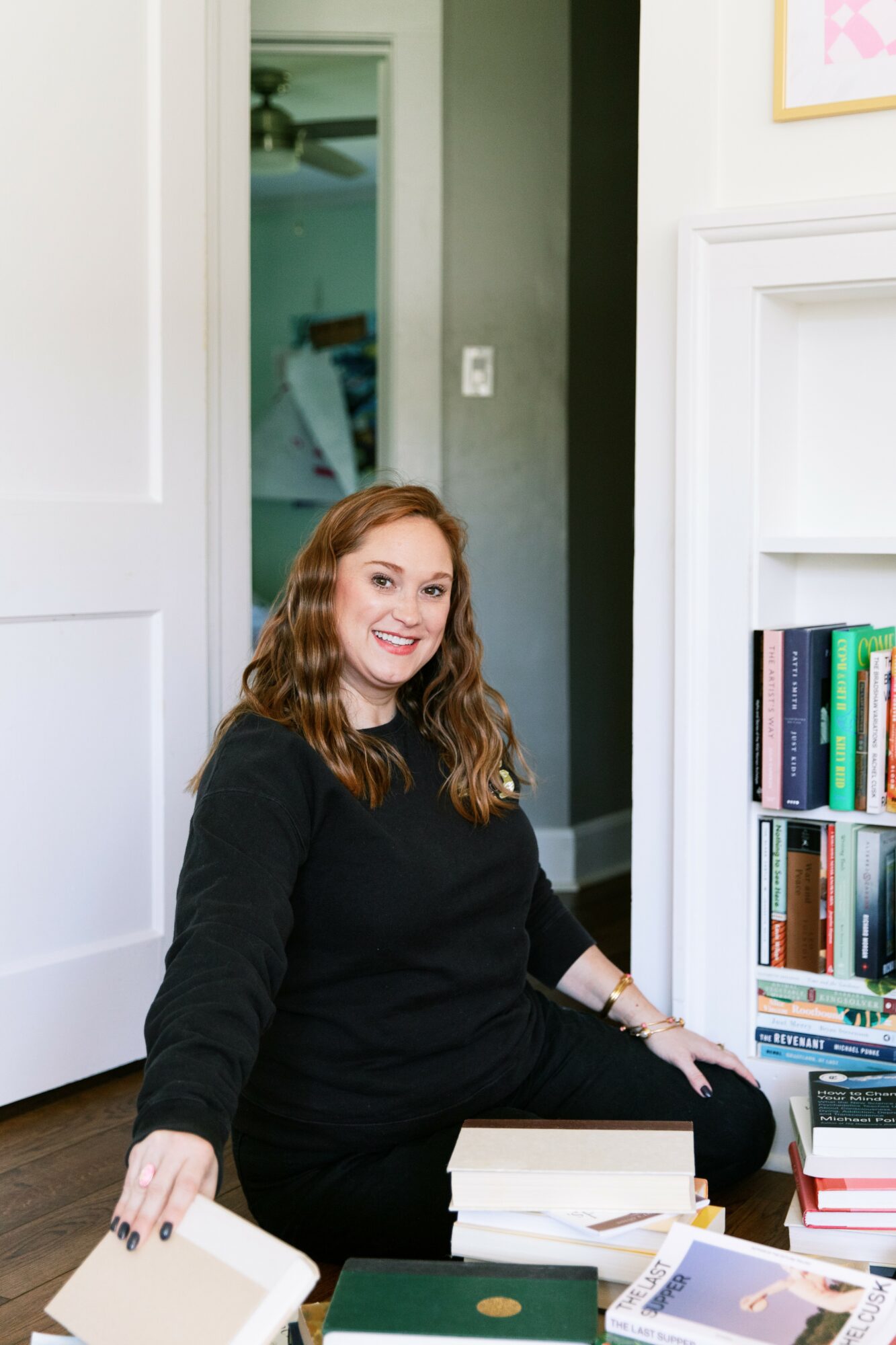 Woman with curly hair sitting on the floor next to a bookshelf, smiling, in a room with white walls and doorways.