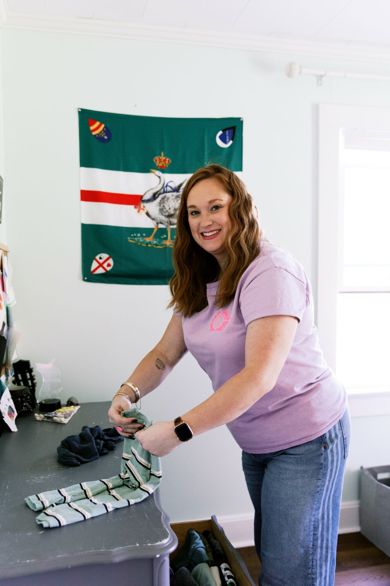 Woman with wavy hair smiling, wearing a light purple shirt, standing in a room with a flag on the wall, folding clothes.