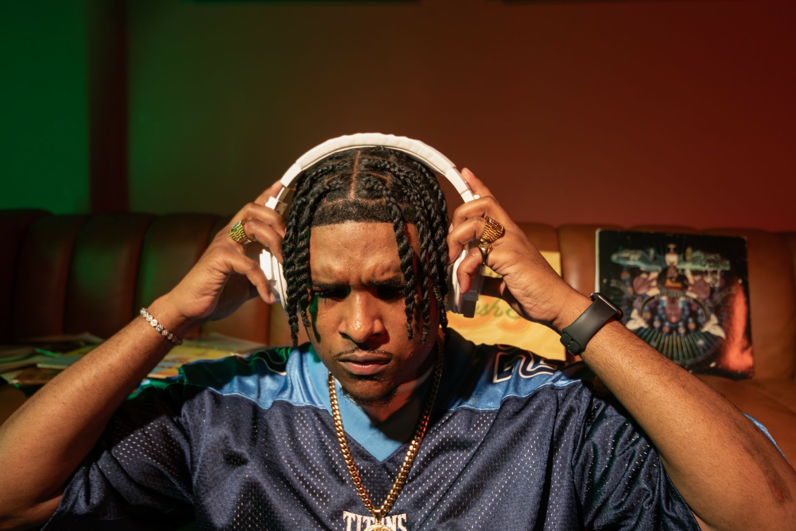 Person with braided hair wearing headphones, a sports jersey, and jewelry, adjusting headphones indoors.