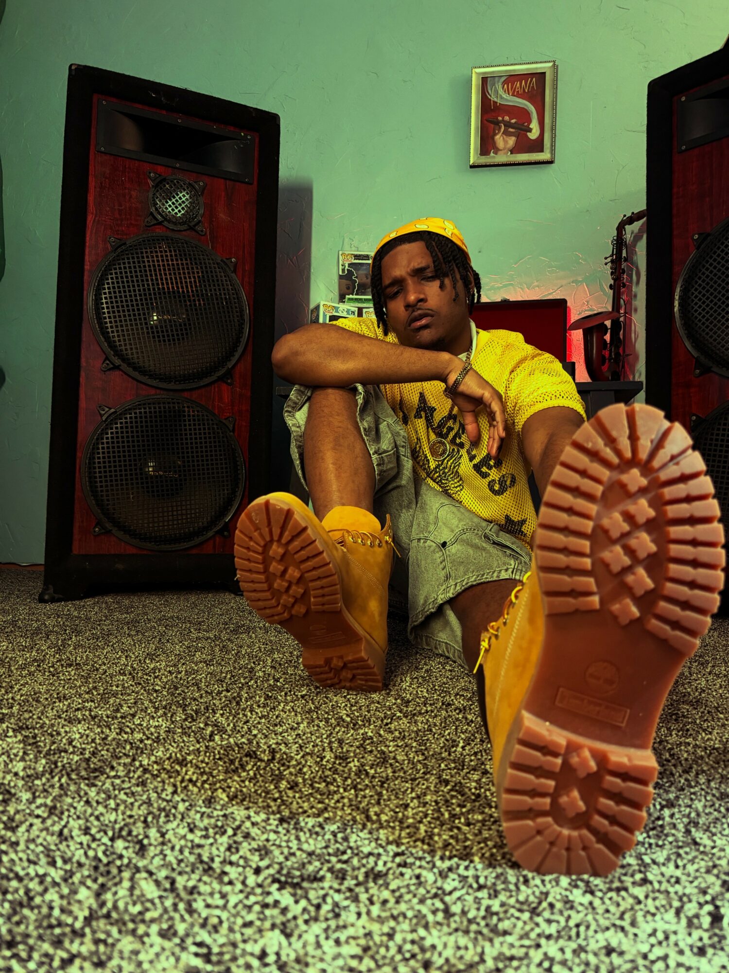 Young person sitting on the floor between two large speakers, wearing yellow clothing and yellow boots, with a green wall background.