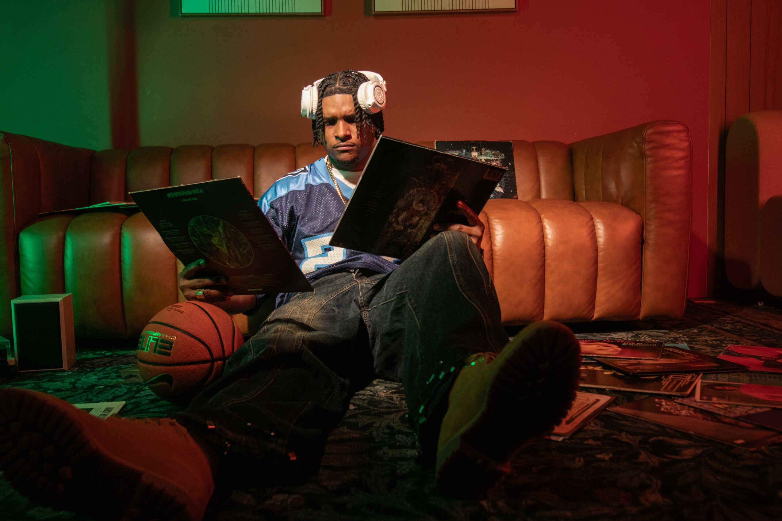 Person sitting on a couch with headphones, reading a magazine, with a basketball and speaker nearby, in a dimly lit room.