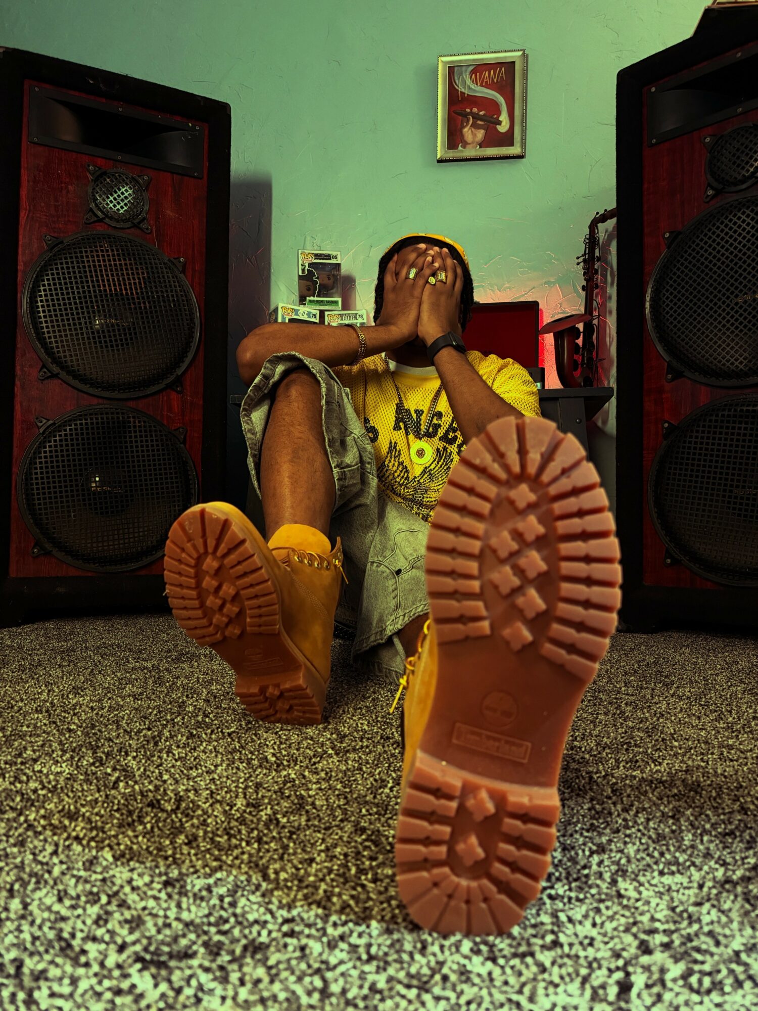 Person sitting on the floor with hands over face, large yellow boots, speakers on each side, green wall, framed picture above.